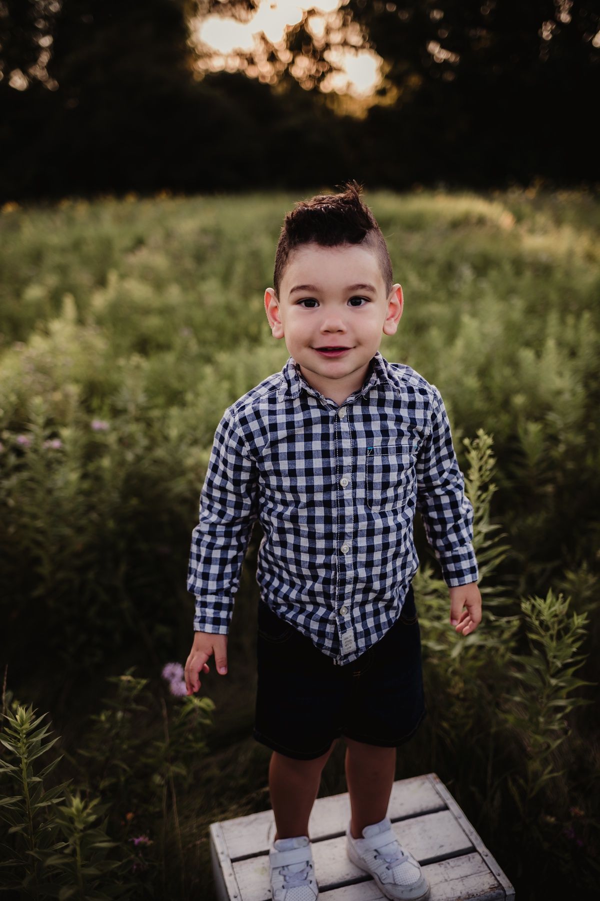 Young child standing on wooden platform wearing checkered shirt and shorts in evening sunlight.