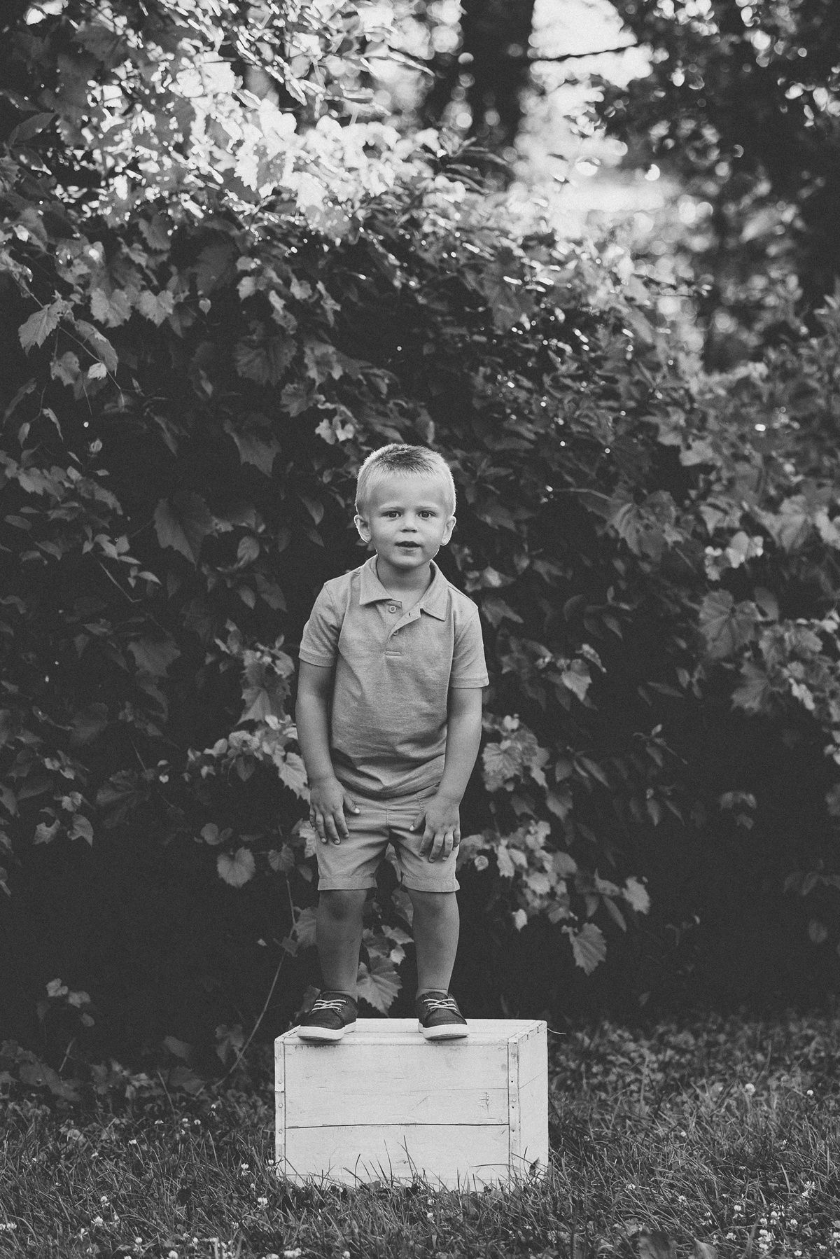A black and white photo of a small child standing on a wooden crate in a garden setting.