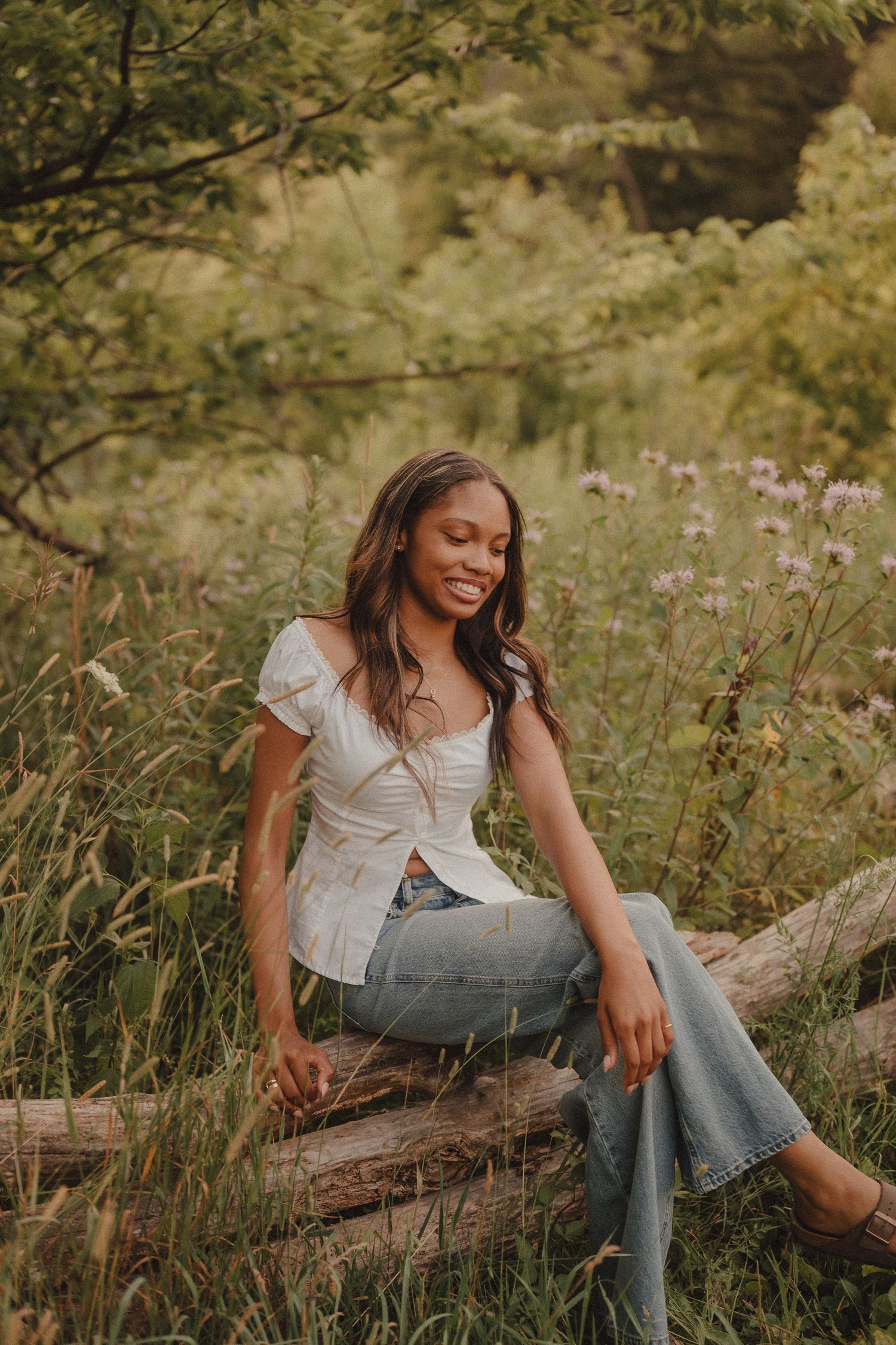 Young woman in white top and jeans sitting on a log in a lush green meadow with wildflowers.