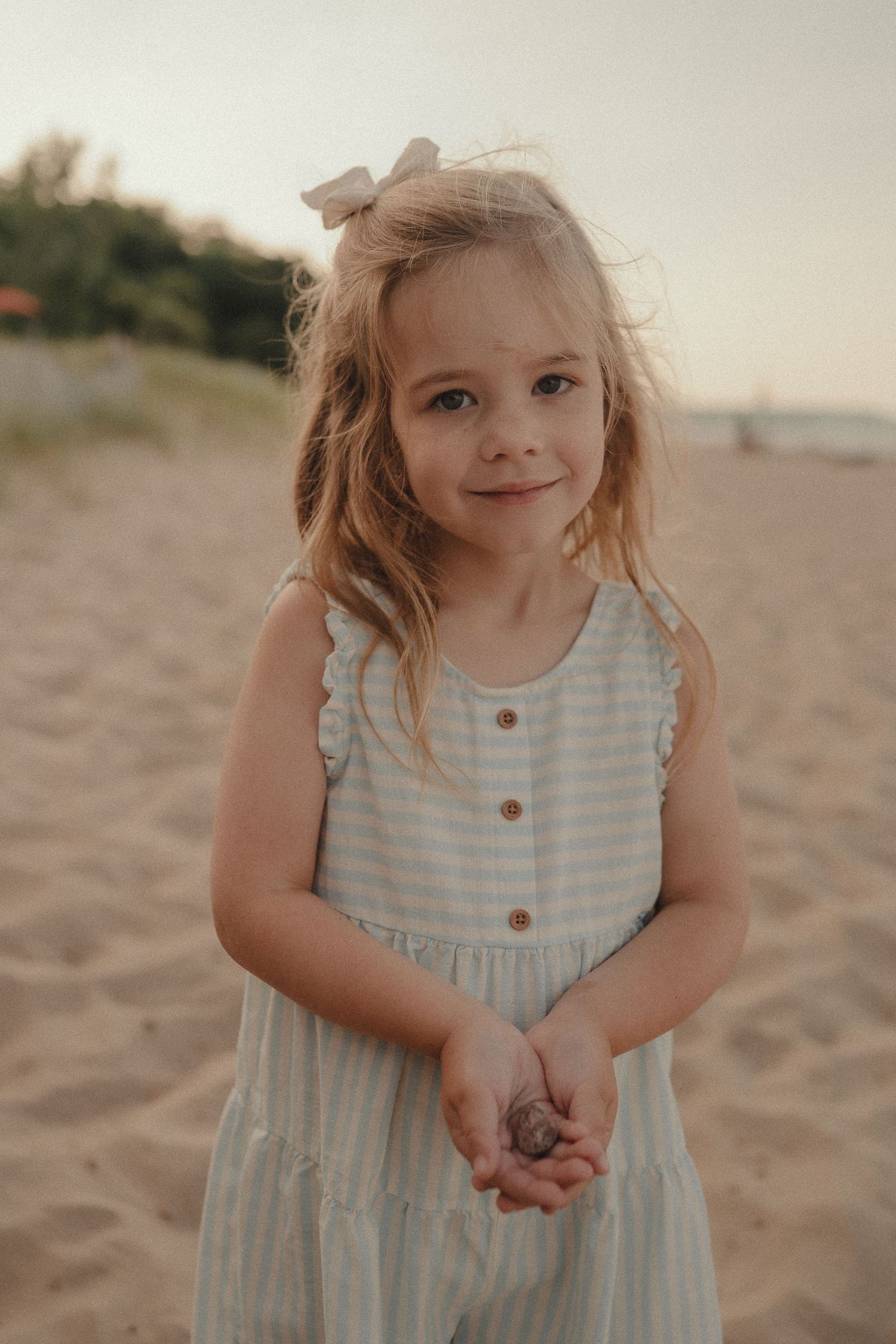Adorable blonde girl in striped dress holding seashells on sandy beach at sunset.