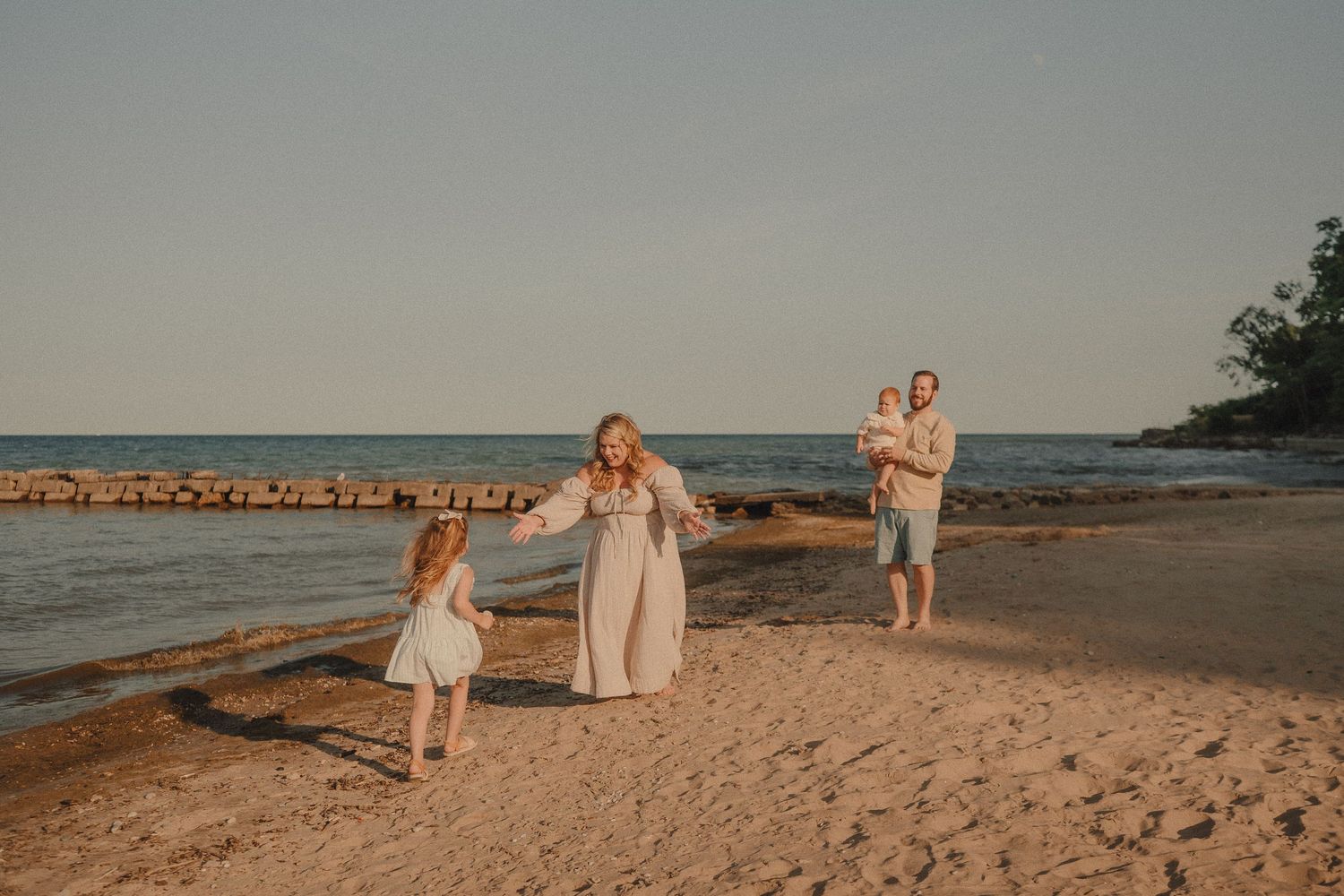 Family of four enjoying a golden hour beach walk, mother twirling daughter on sandy shore near calm water.