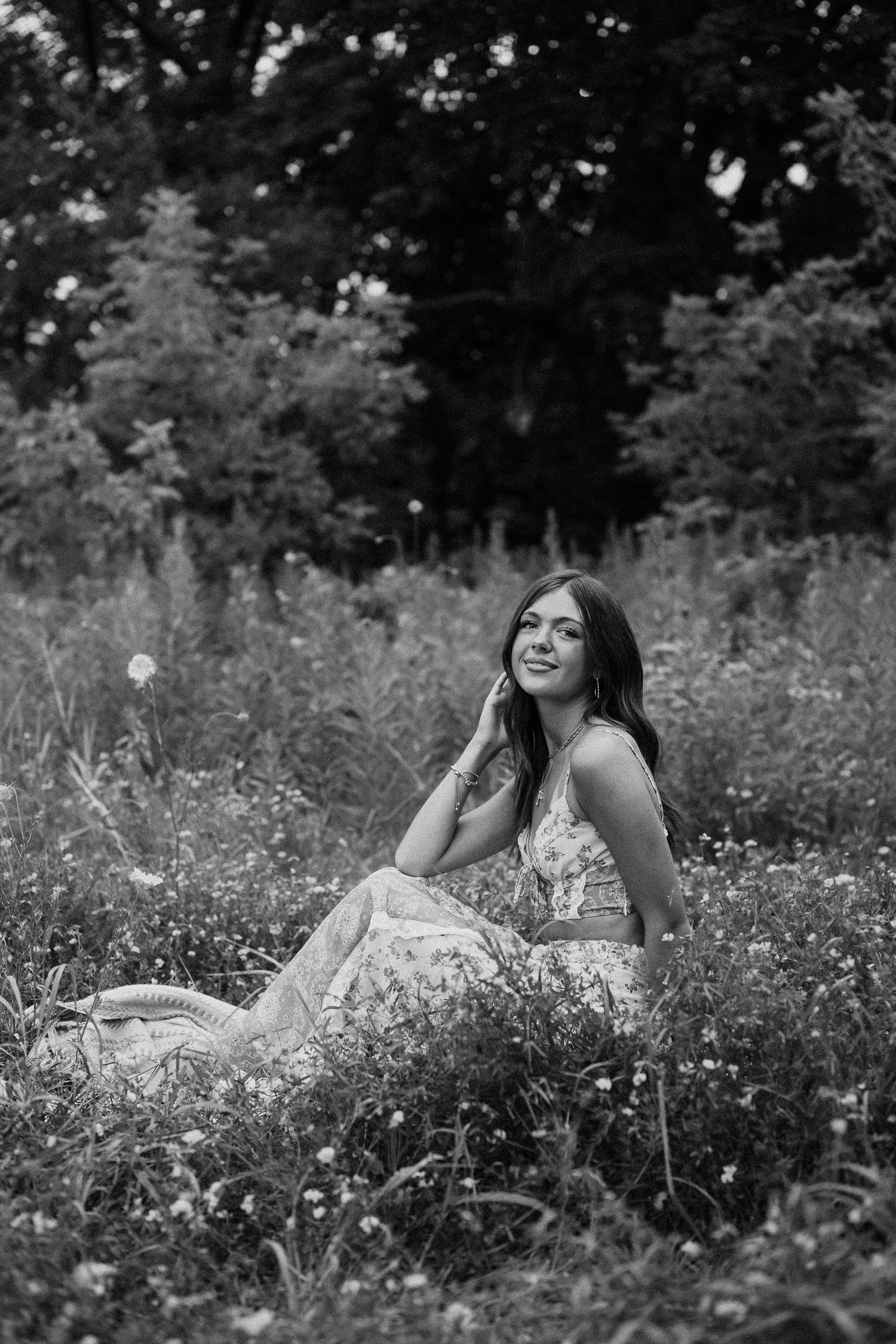 Black and white photo of a smiling young woman sitting in a wildflower field touching her face.