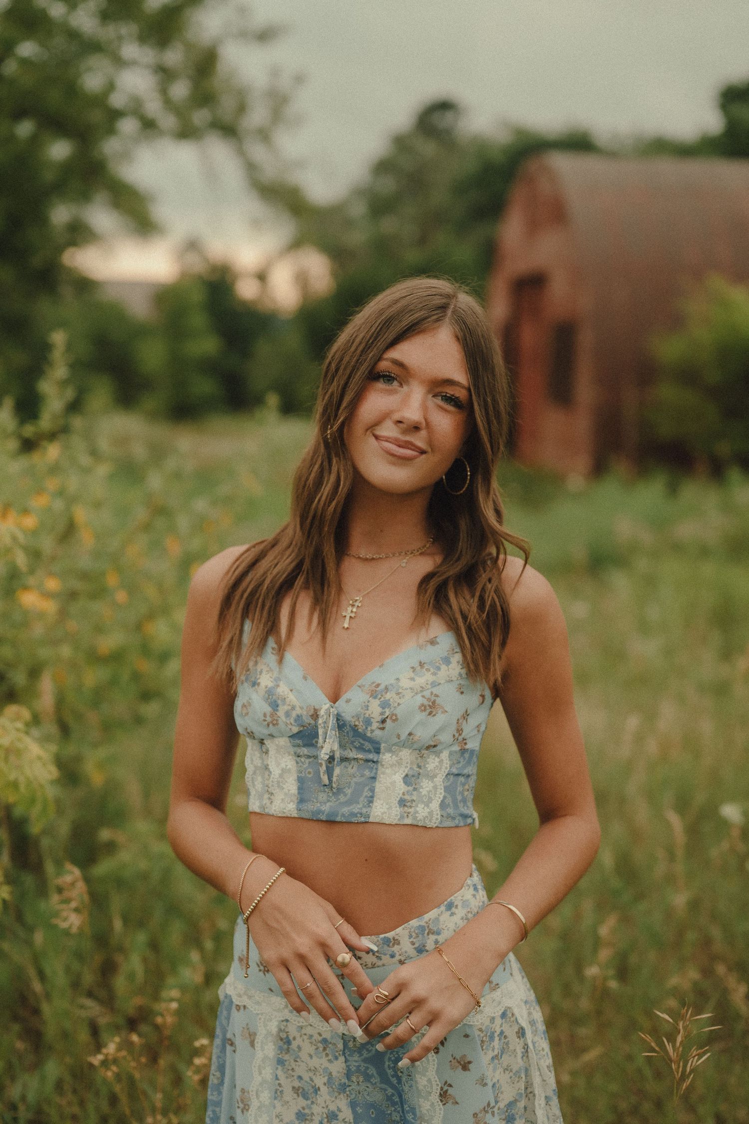 Young woman in floral crop top and skirt smiling in a wildflower field with rustic barn backdrop.