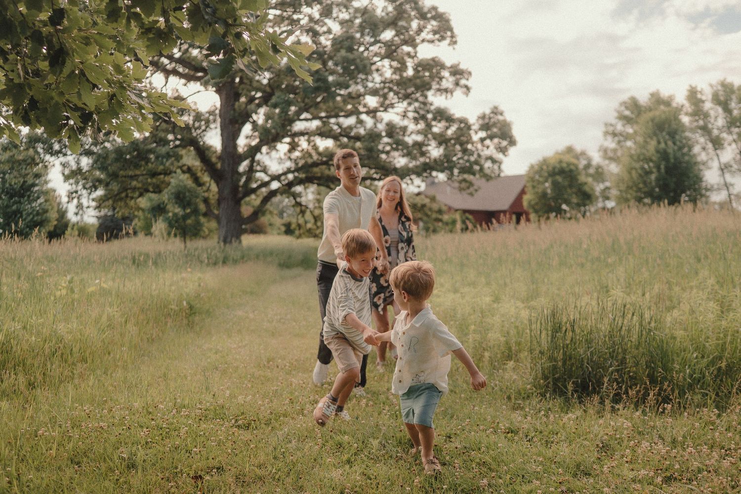 Family with two young children playing in a green meadow near a large oak tree on a farm.