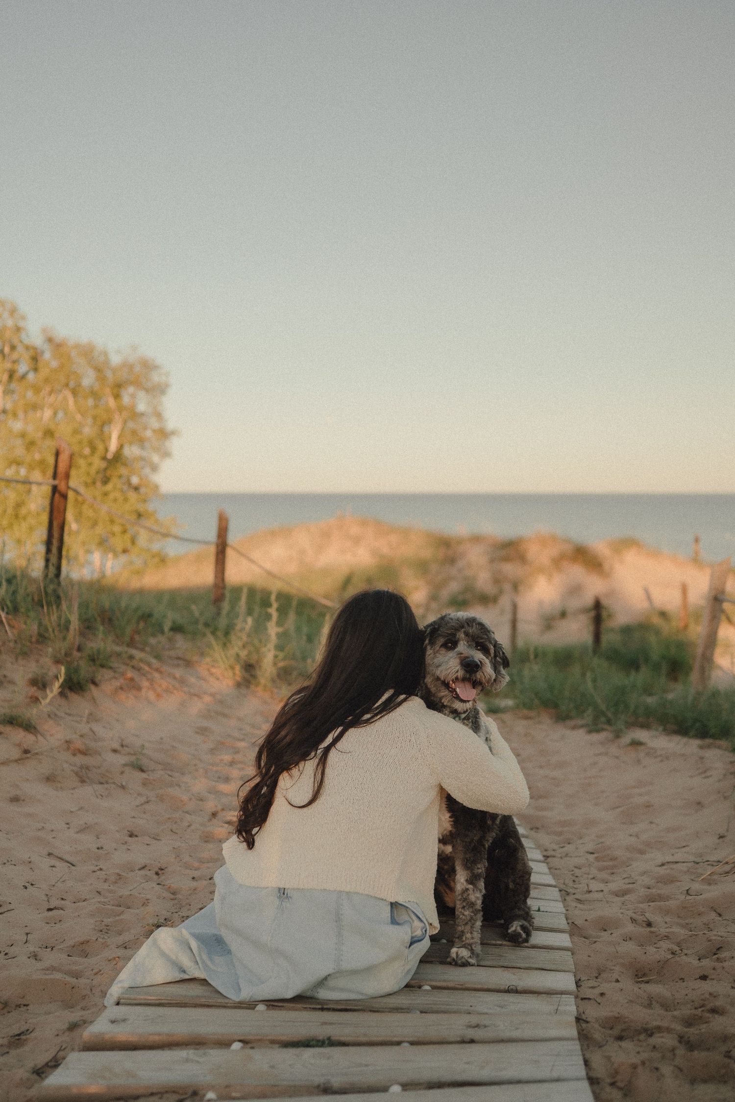 A young woman in a white sweater sits on a wooden boardwalk hugging her fluffy dog near a sandy beach at sunset.