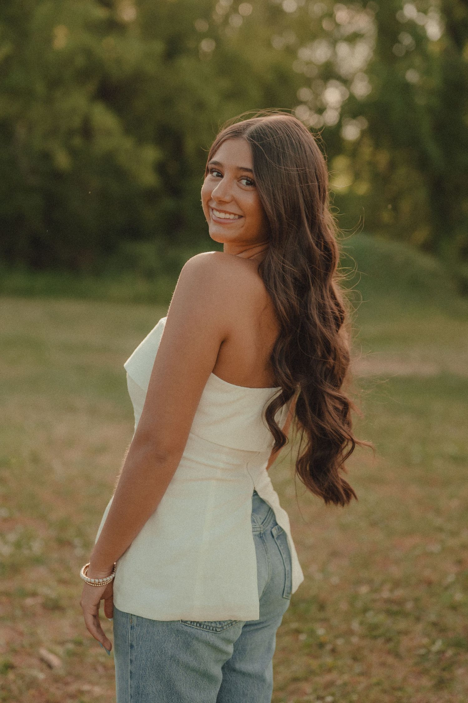 Young woman in white strapless top and jeans smiling over shoulder outdoors at golden hour.