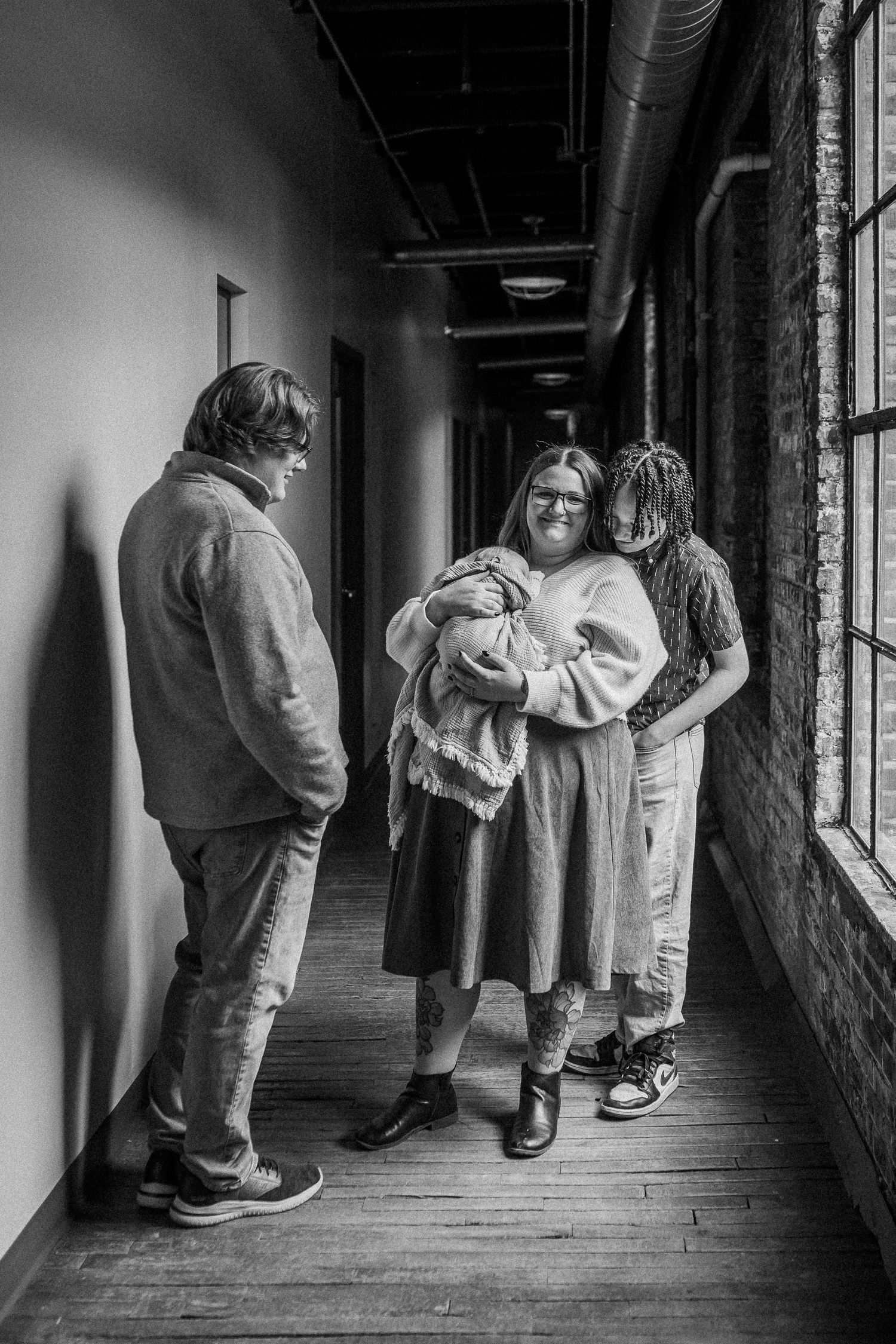 Black and white photo of three people in a hallway, one holding a newborn baby, sharing a tender moment.