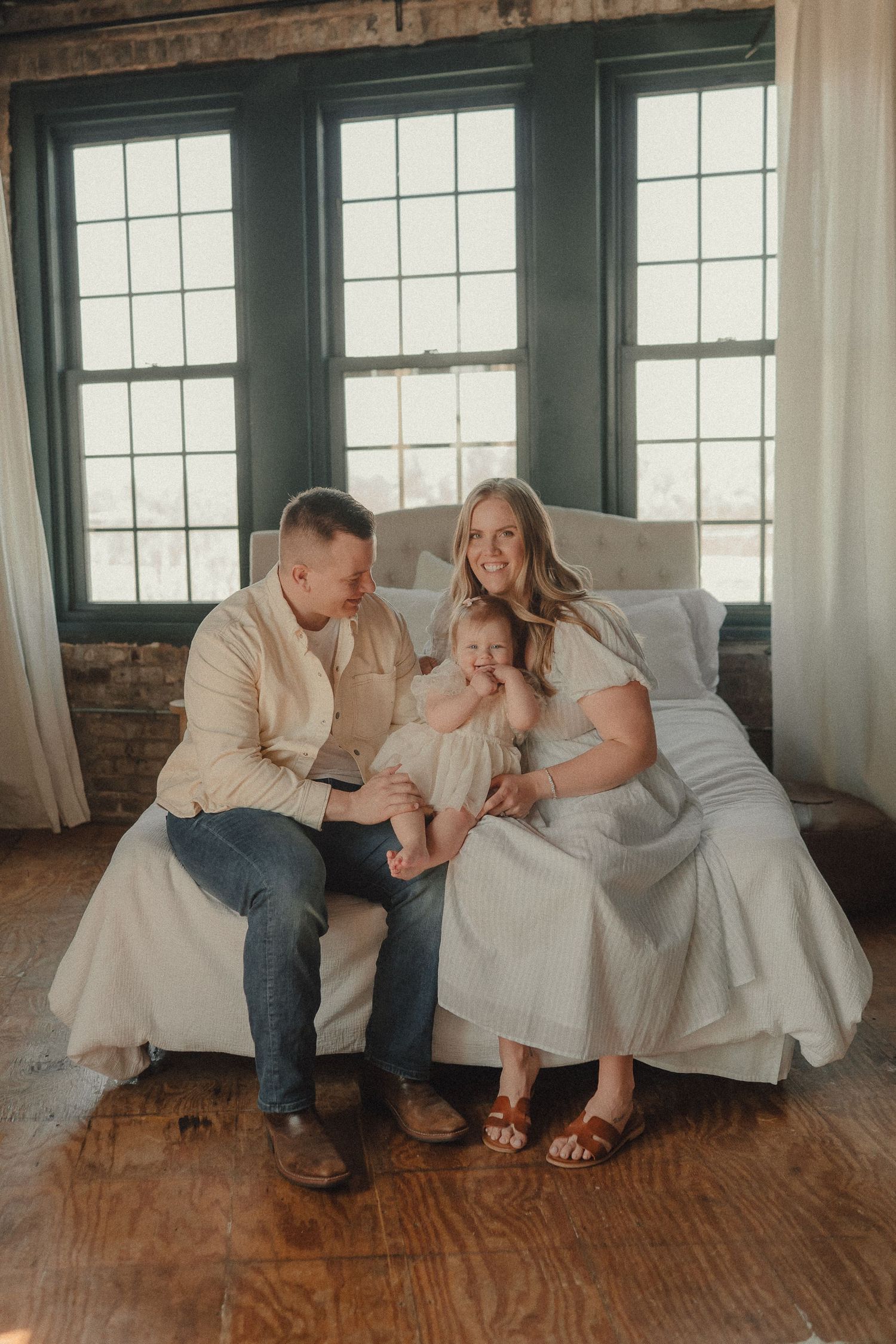 A family of three sits together on a couch near large windows in a warmly lit rustic indoor setting.