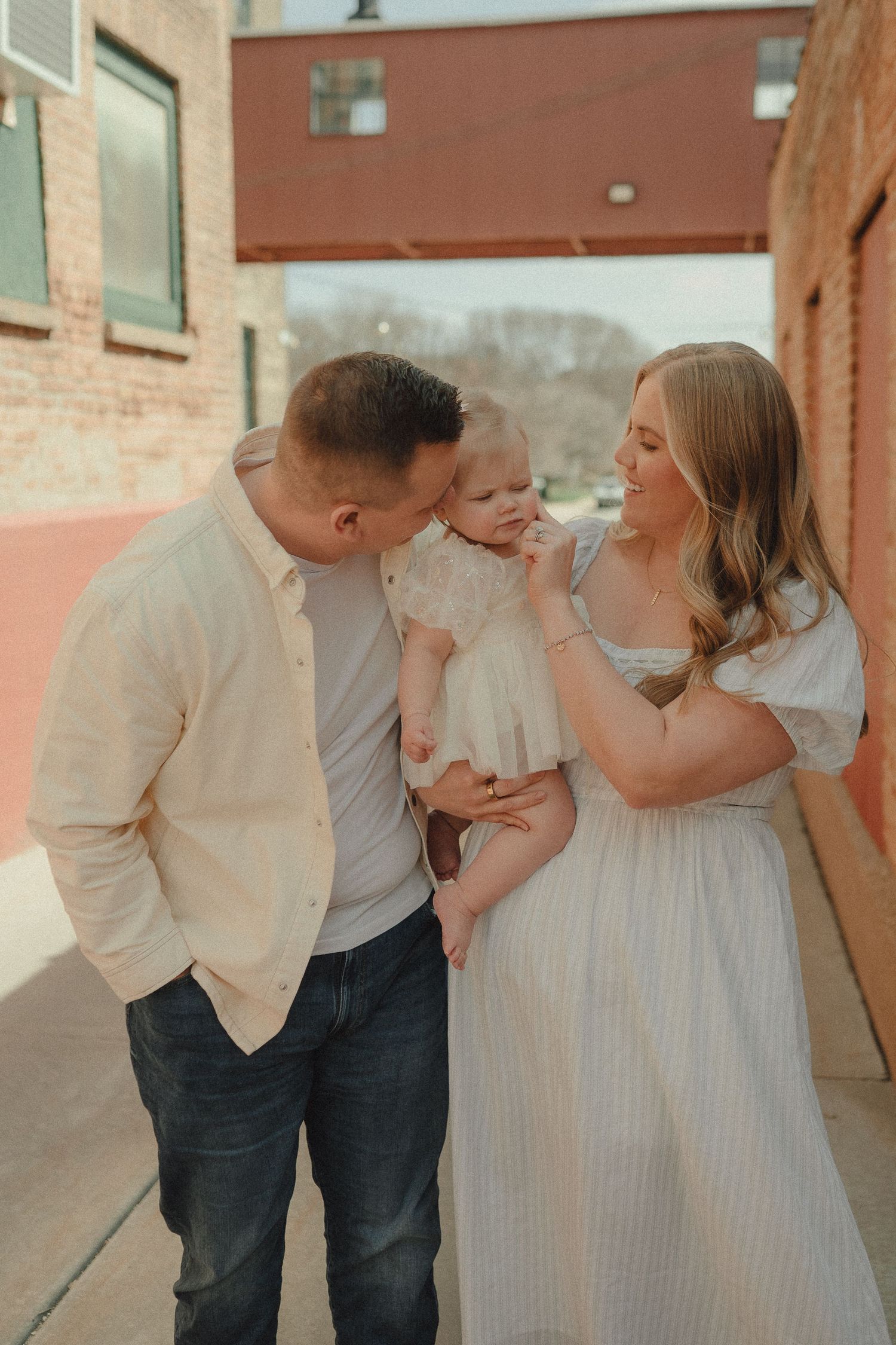 A happy family of three poses outdoors, father kissing baby girl while mother smiles in a light striped dress.