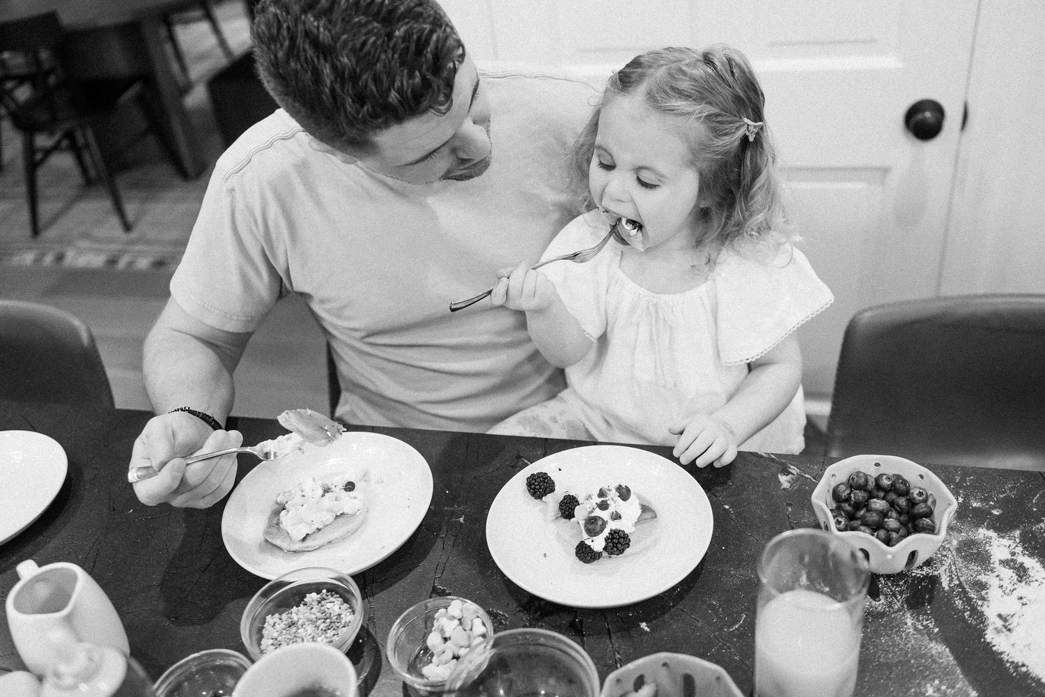 Father feeding his young curly-haired daughter a bite of food at a breakfast table in black and white.
