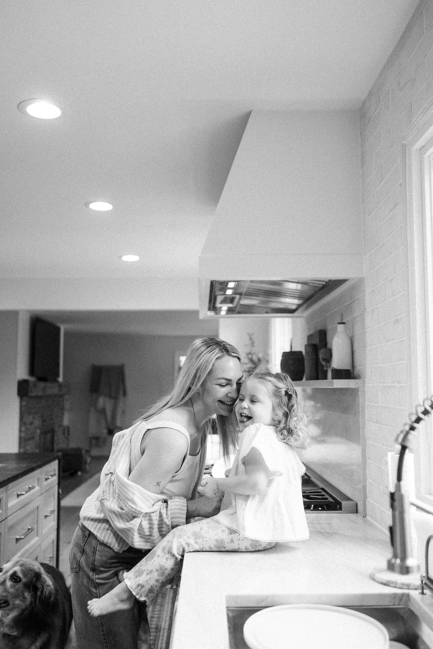 Black and white photo of a smiling mother holding her laughing toddler daughter in a modern kitchen.