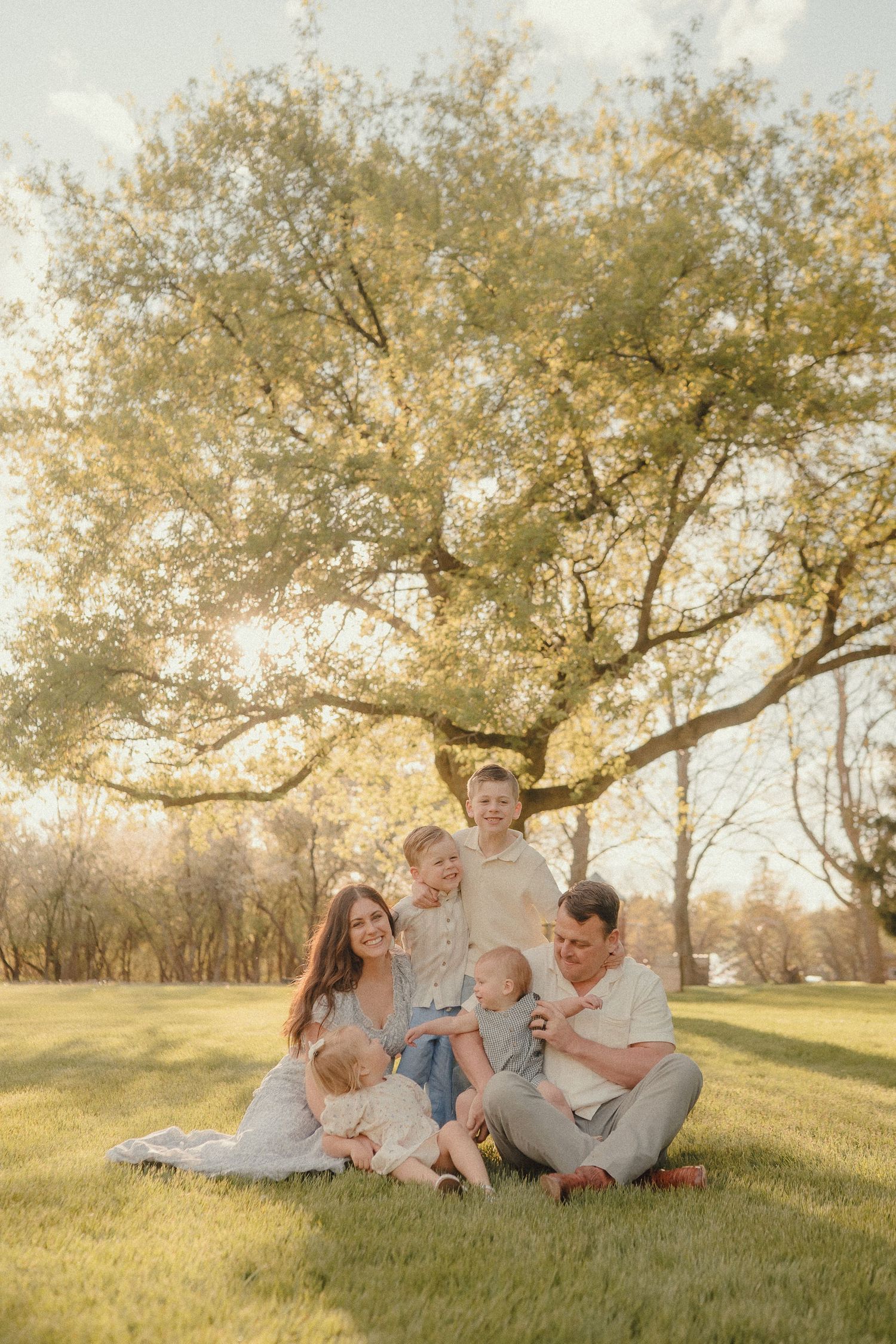 Family of six sitting on grass under a large tree in golden hour light, wearing neutral tones.