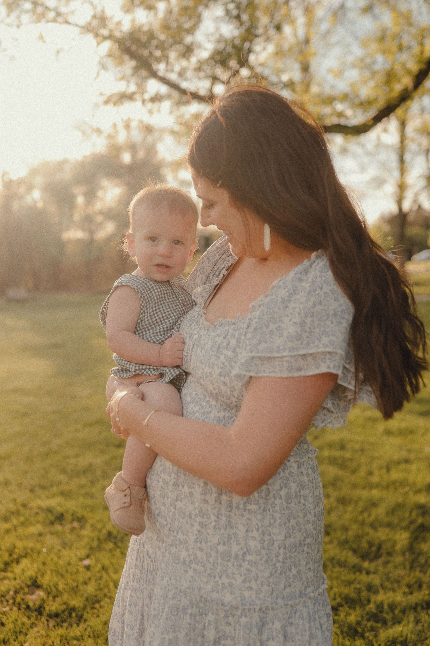 Mother in floral dress tenderly holding her baby outdoors in warm golden sunset light near trees.