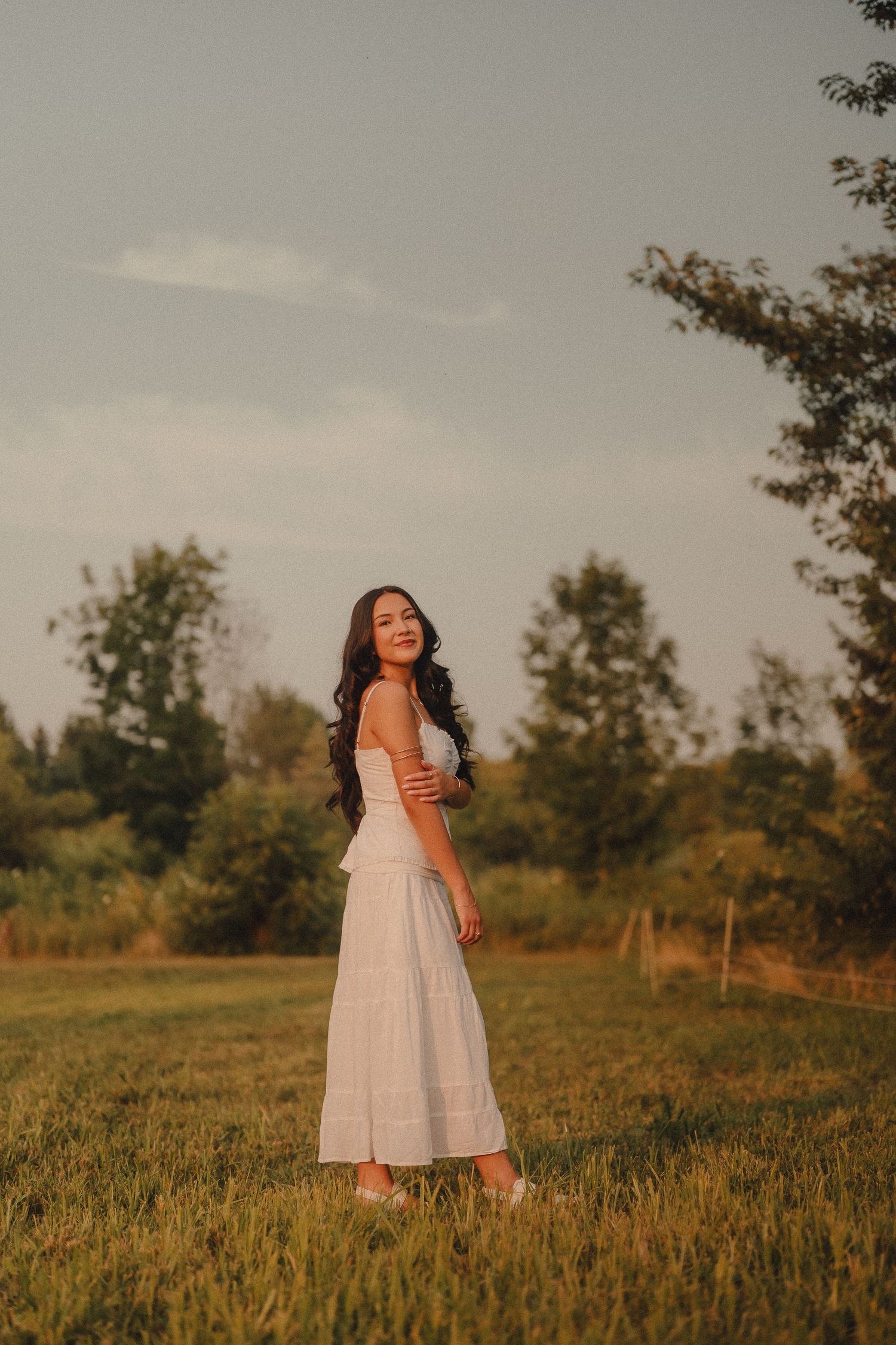 Smiling woman in white dress standing in green meadow during golden hour sunset.