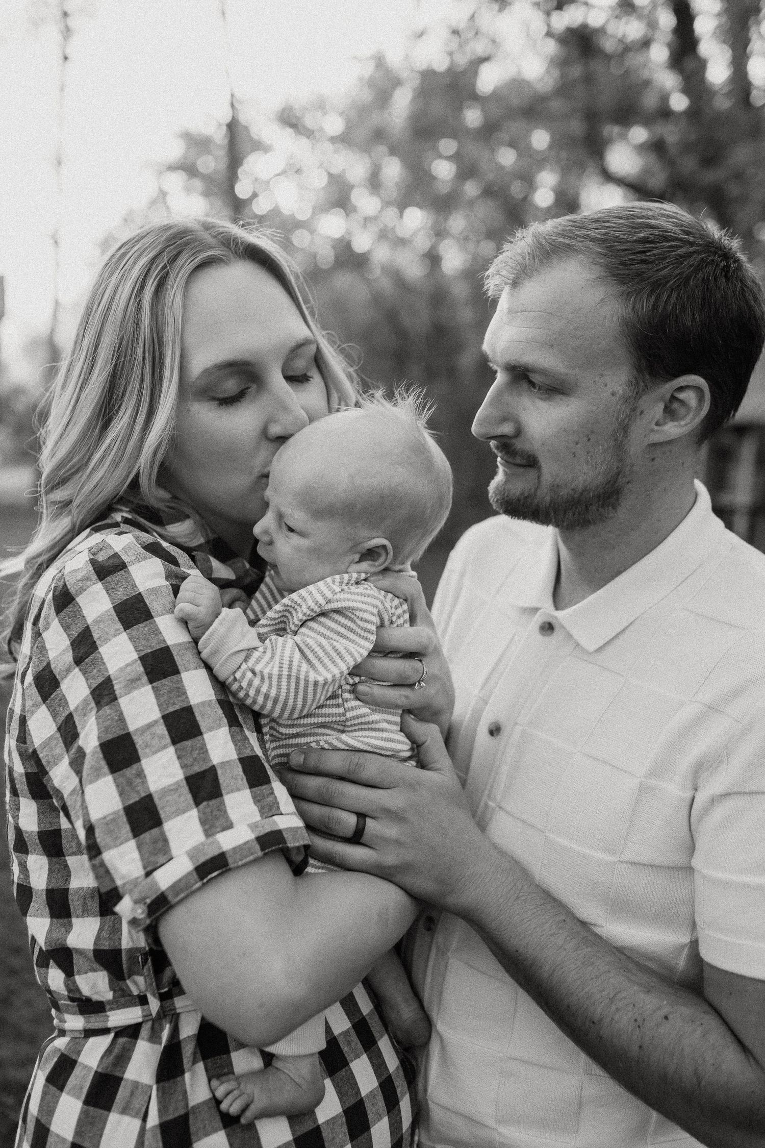 Loving parents in black and white photo, mother kissing newborn baby held by father outdoors.