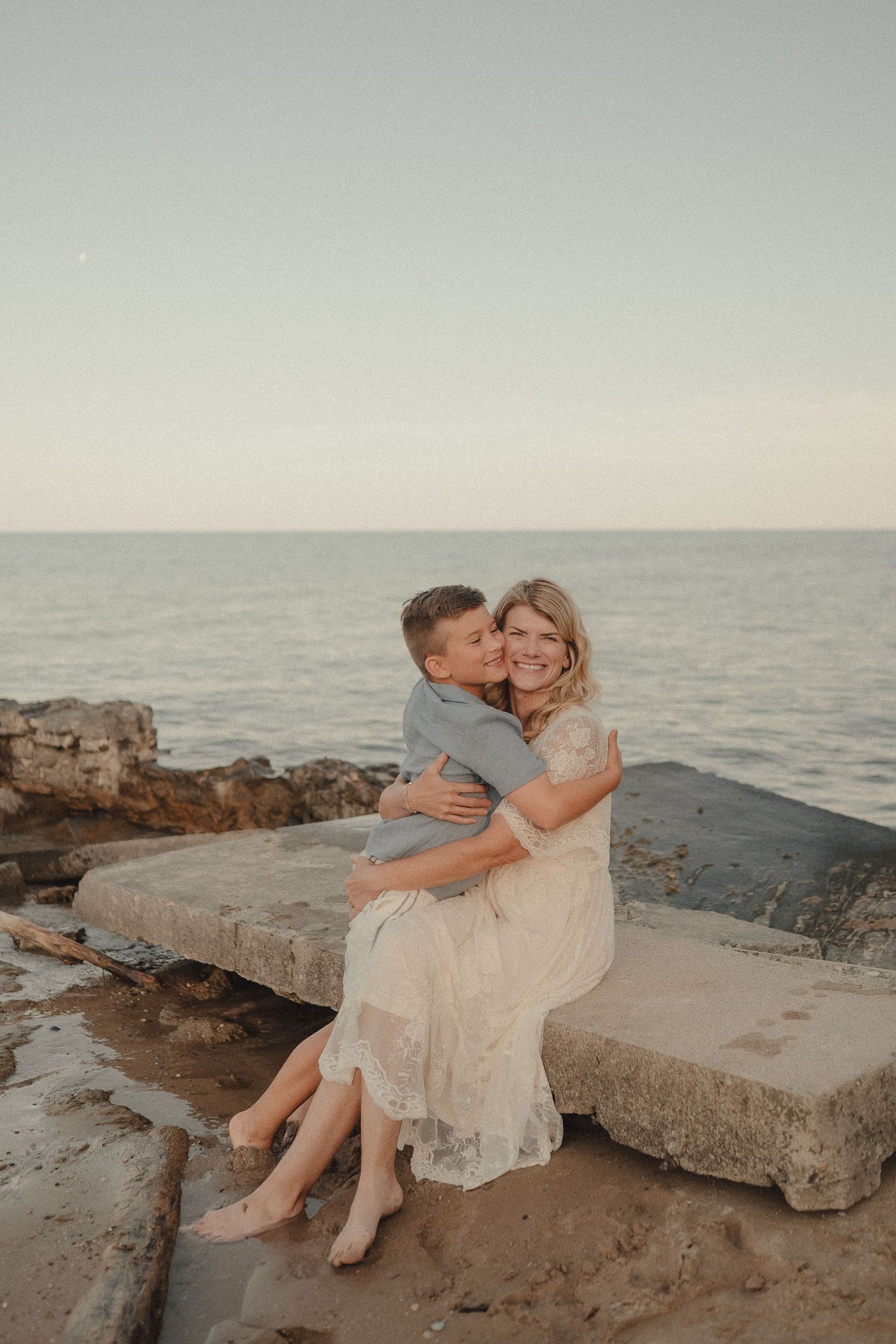 A woman in a white lace dress and a boy share a warm hug while sitting on a stone ledge by the ocean.