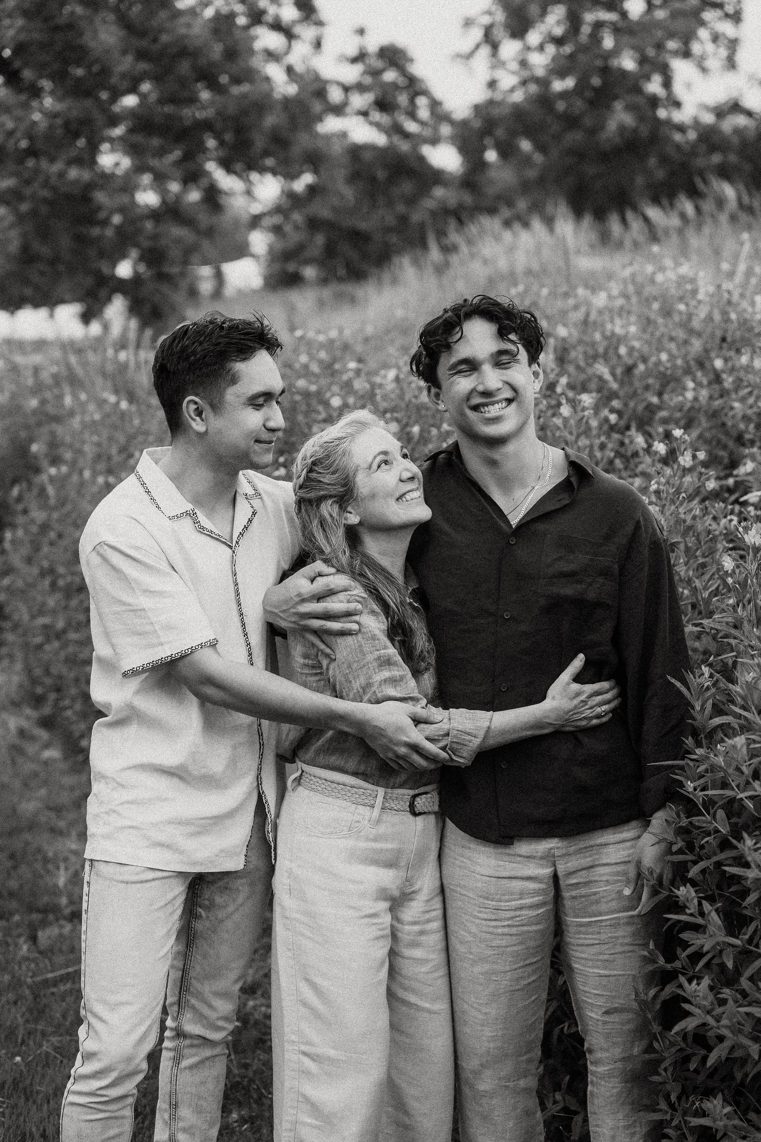 Black and white portrait of young woman embraced by two young men in garden setting.