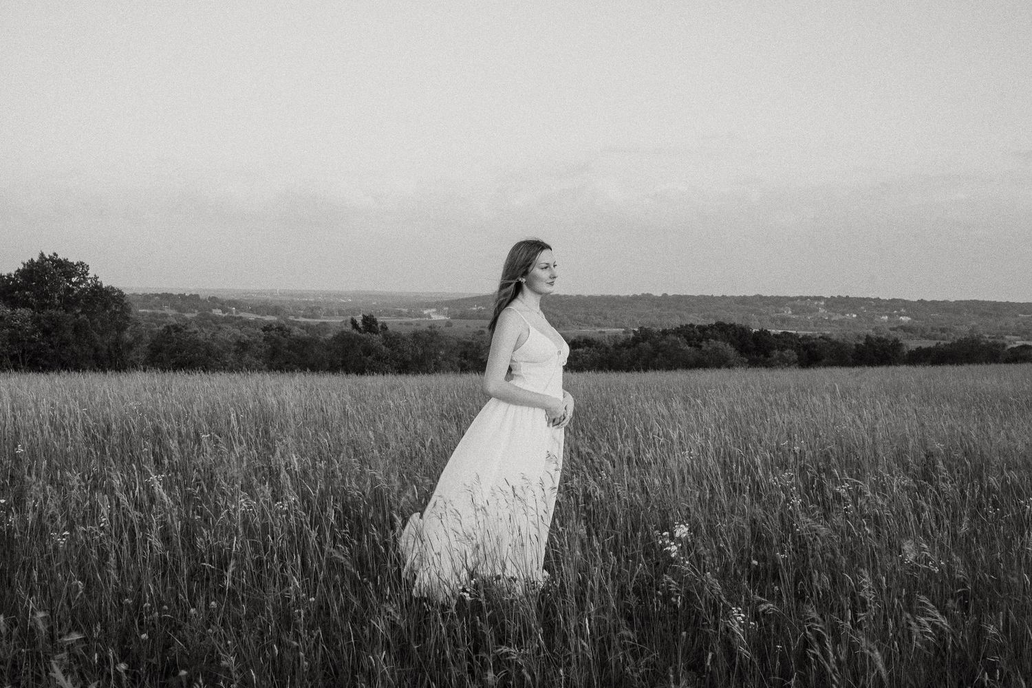 Black and white photo of a woman in a white dress standing in a lush field under a soft sky.