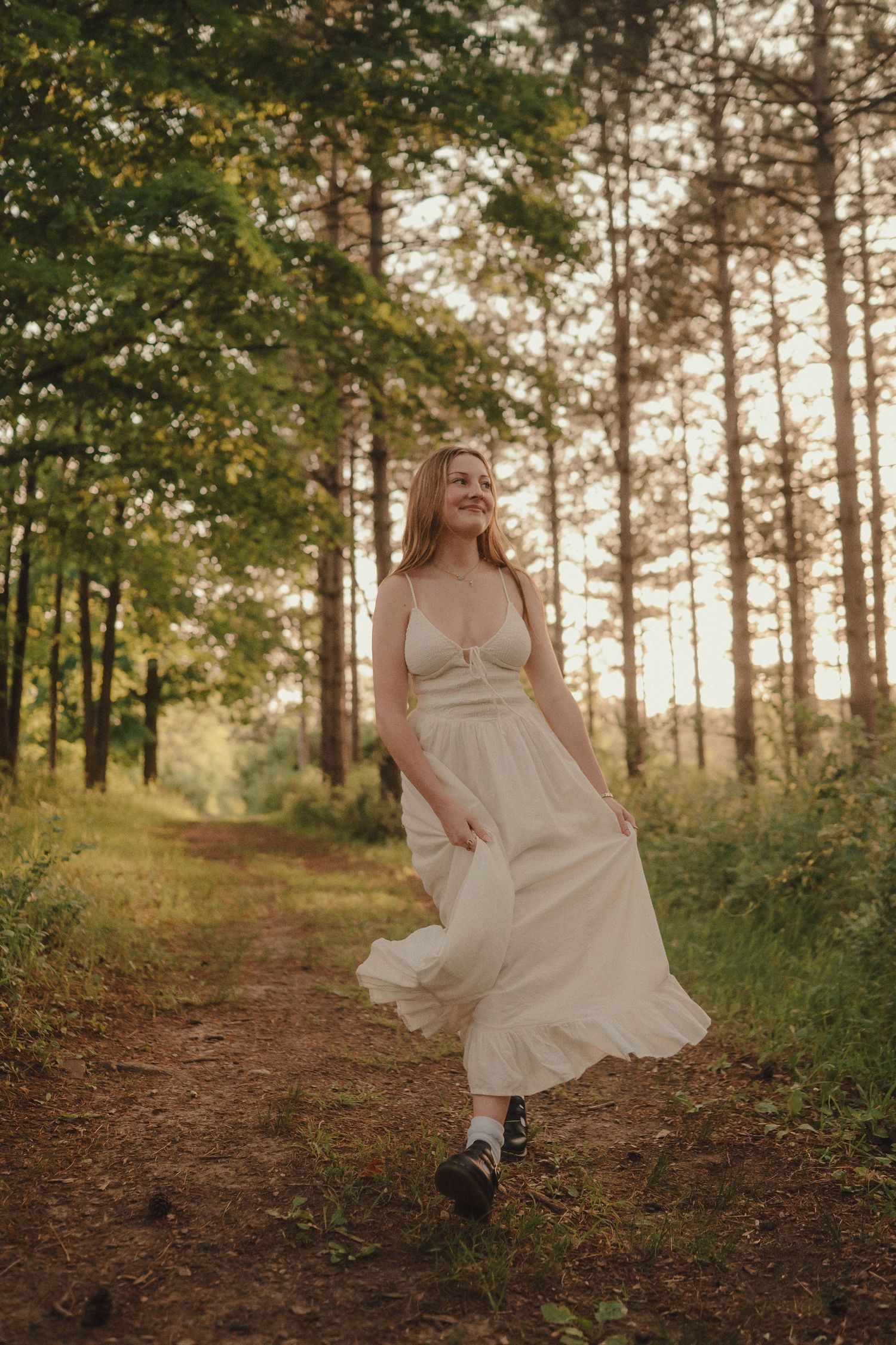 Smiling young woman in a white dress twirls on a sunlit forest path surrounded by tall pine trees.