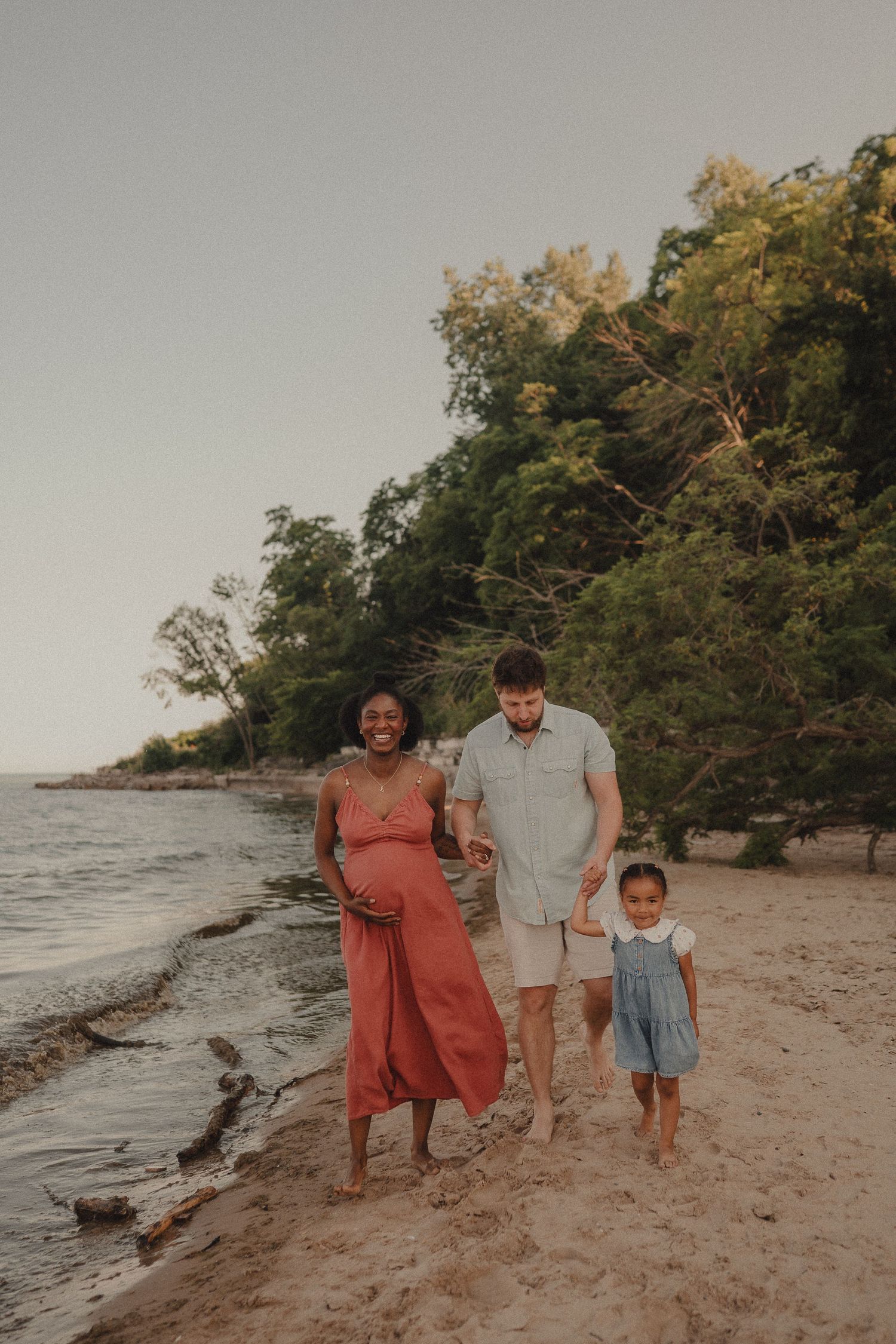 Expecting interracial family walking barefoot on sandy beach at sunset, mother in coral maternity dress.