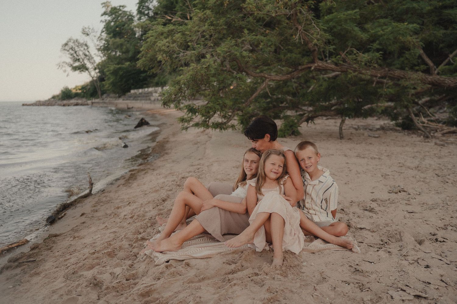 Family sitting together on sandy beach near water surrounded by lush green trees.