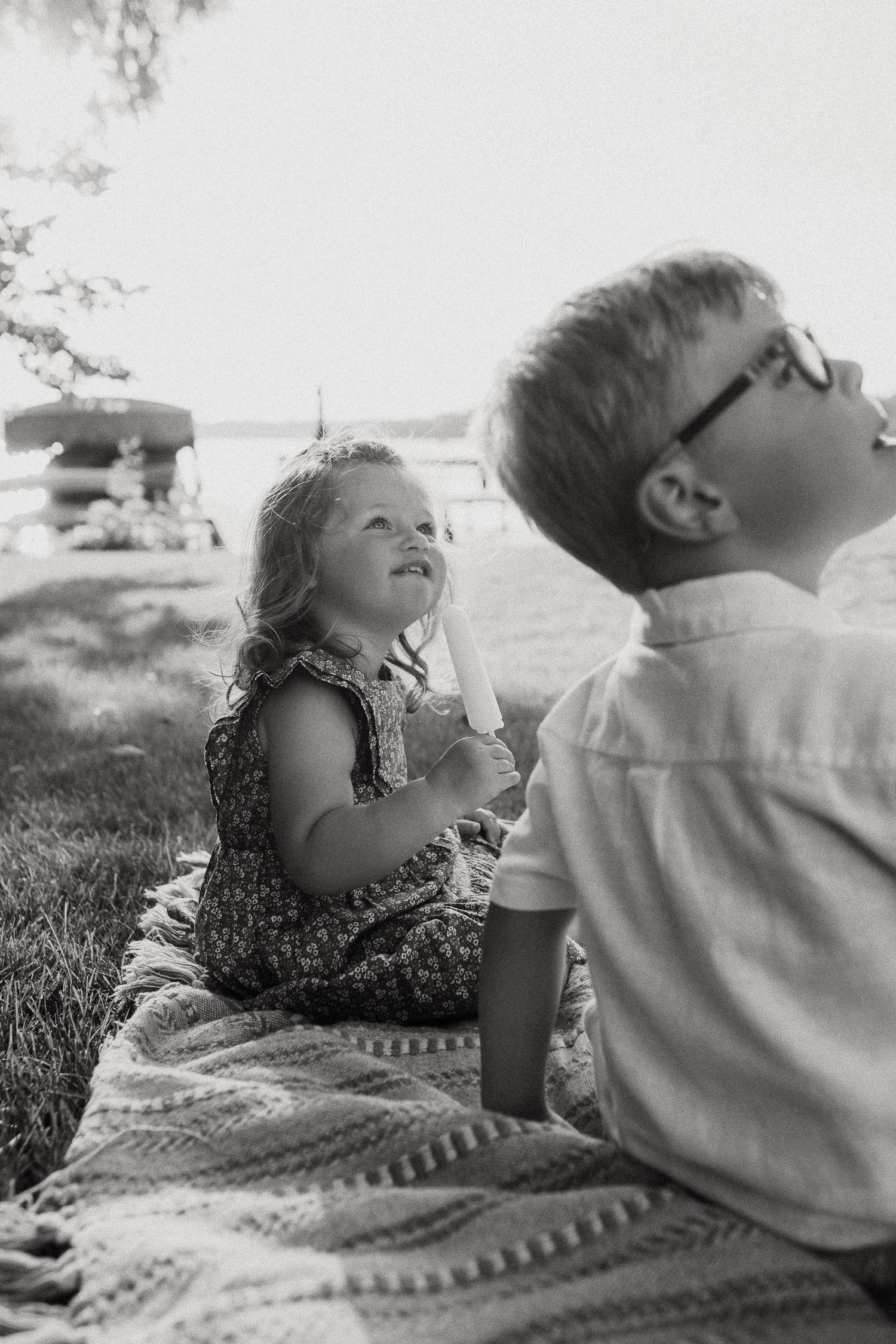 Two children sit on a blanket near a lake, enjoying popsicles on a sunny day.