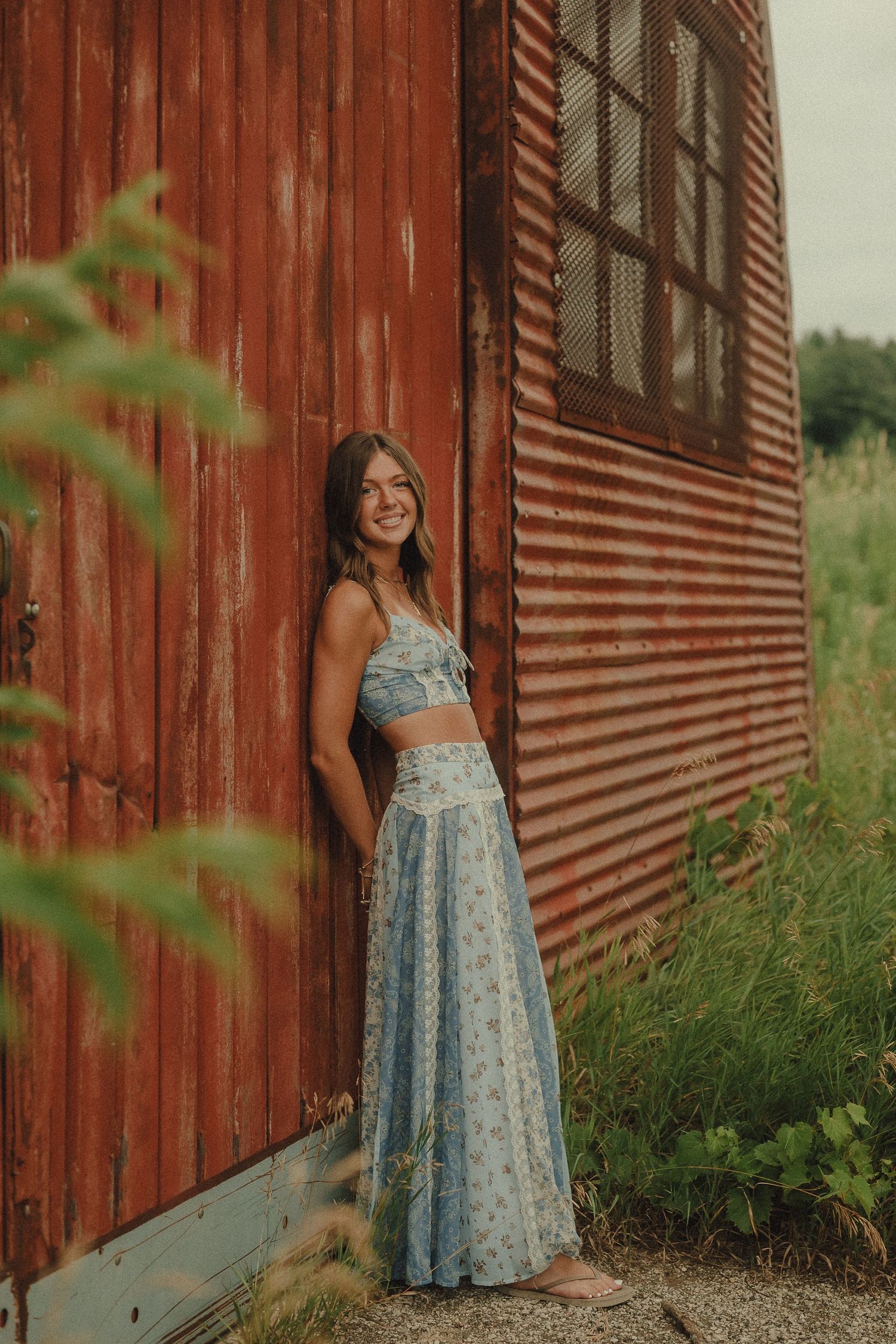 Smiling young woman in blue floral crop top and maxi skirt leaning against rustic red barn outdoors.