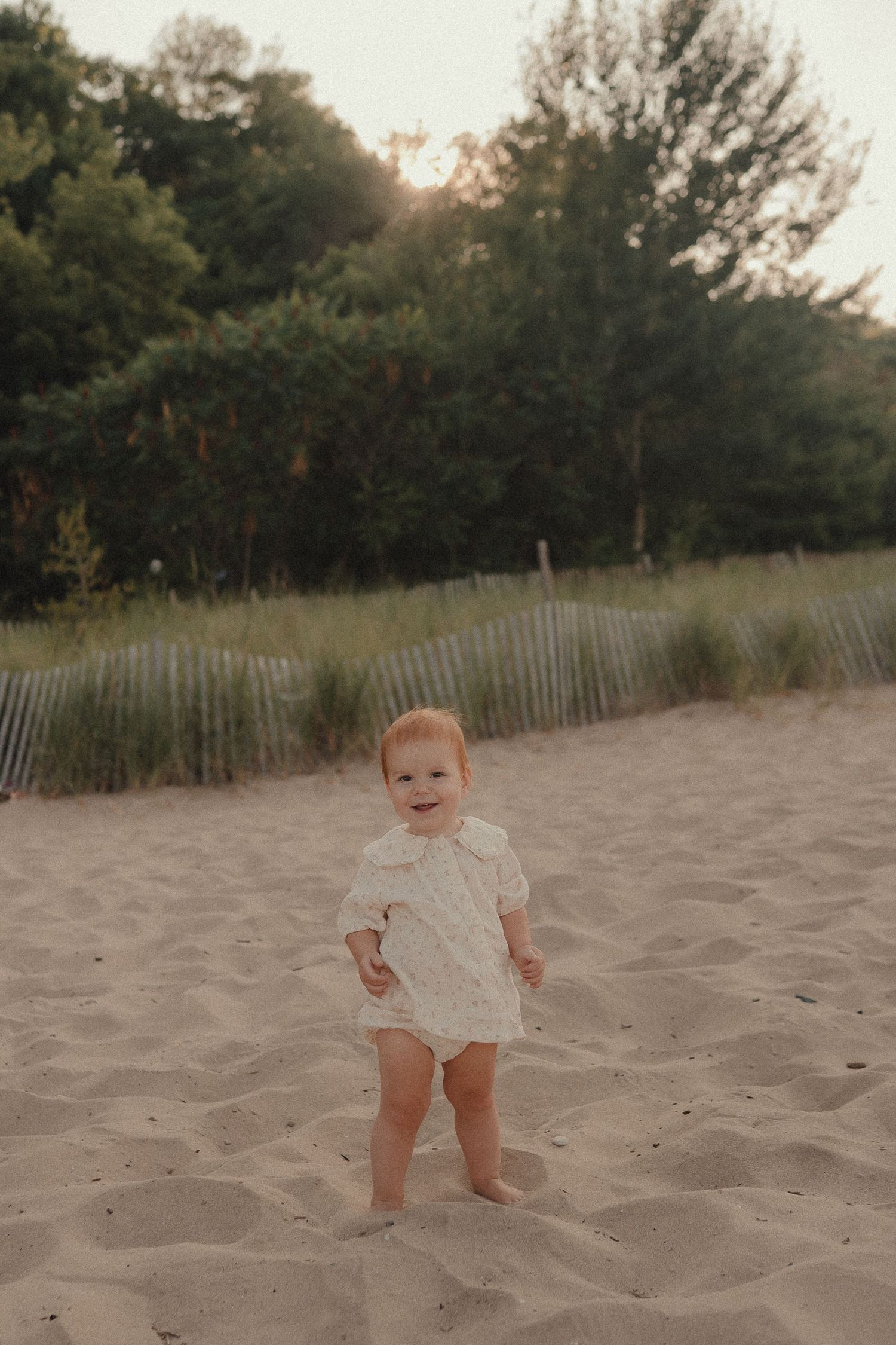 Smiling red-haired toddler in white dress standing on sandy beach at golden hour with trees in background.