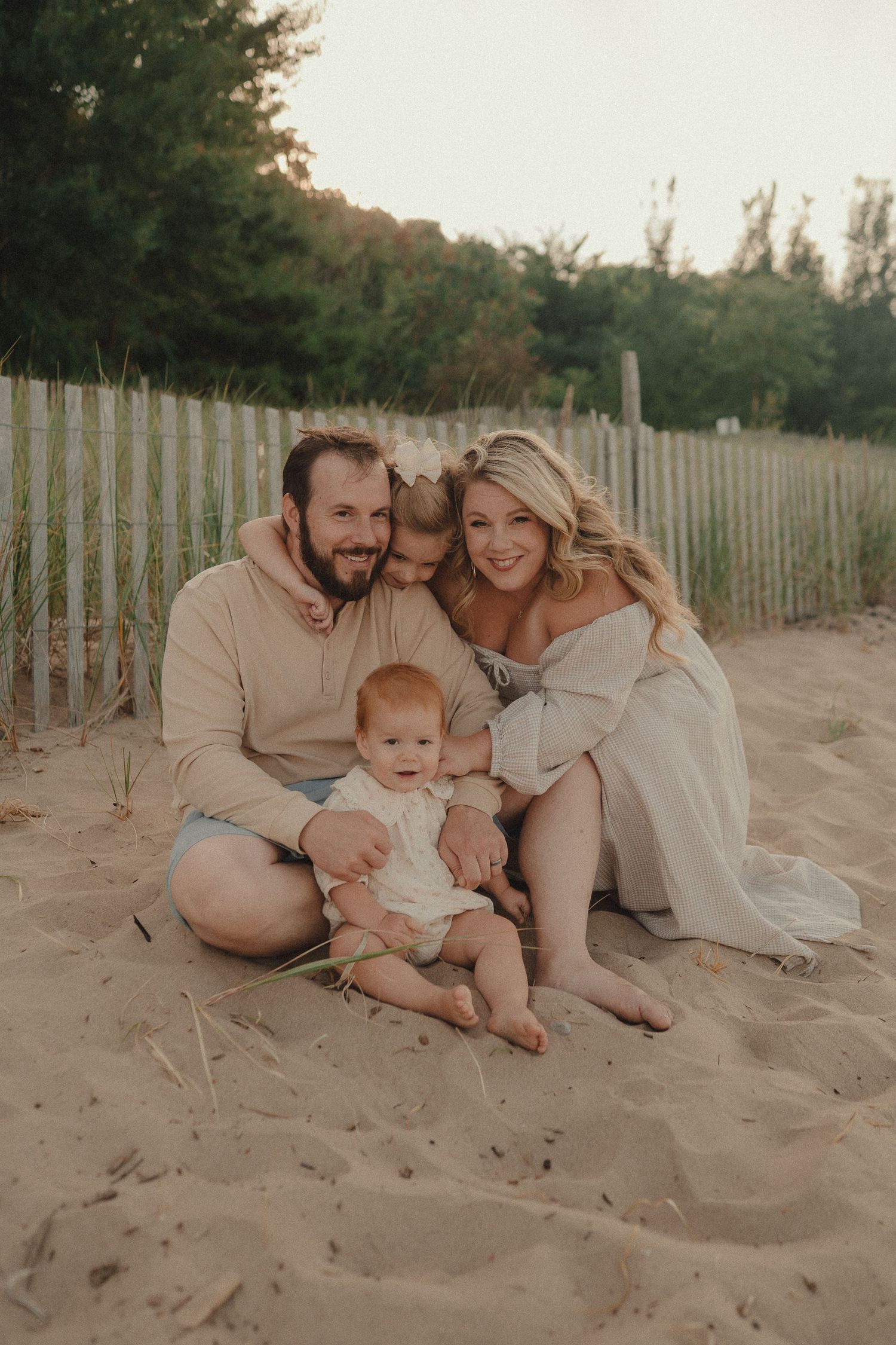 Family of four sitting on sandy beach near wooden fence, parents smiling with toddler and baby girl.