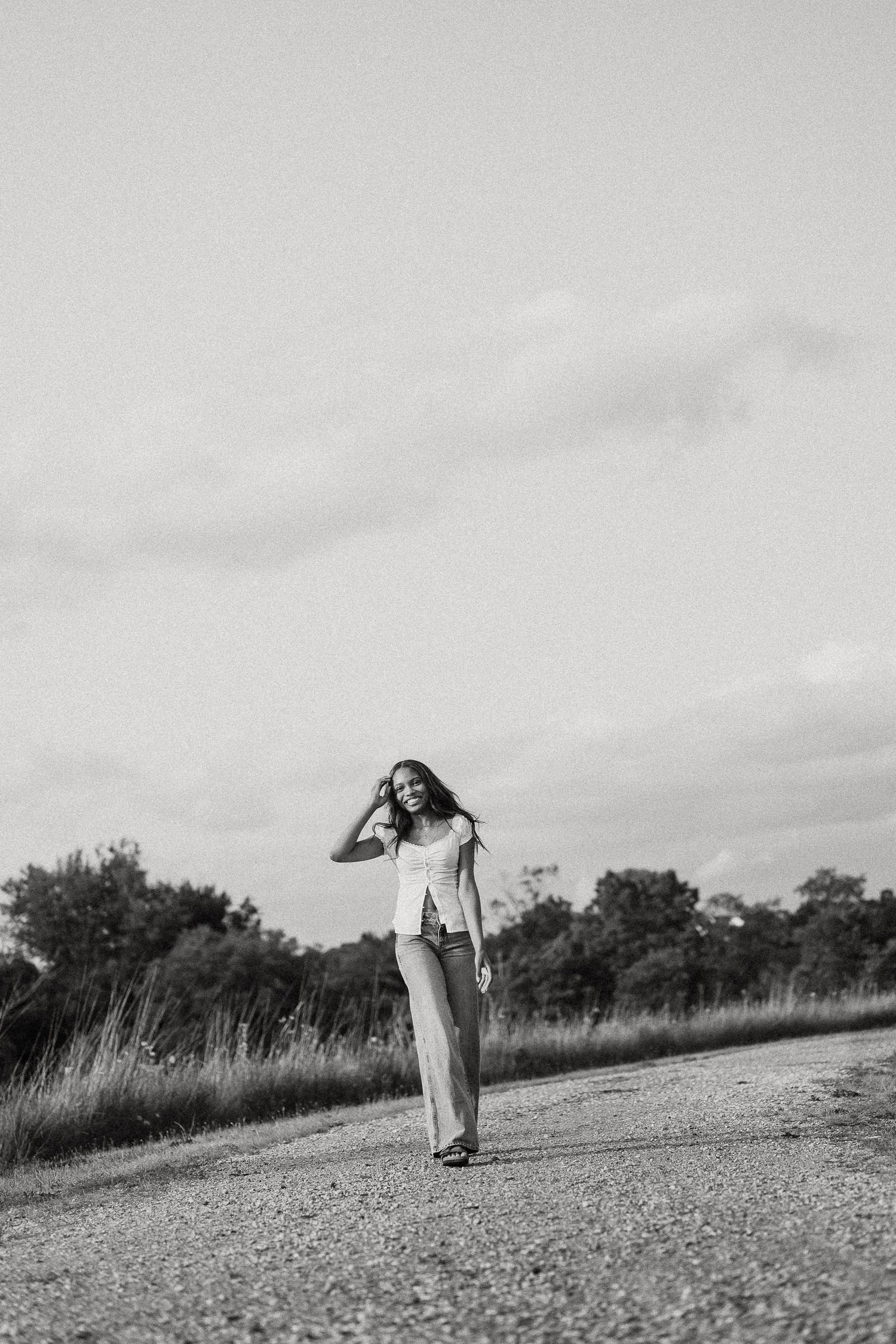 Black and white photo of a young woman walking on a rural road, wearing shorts and a tank top, smiling.