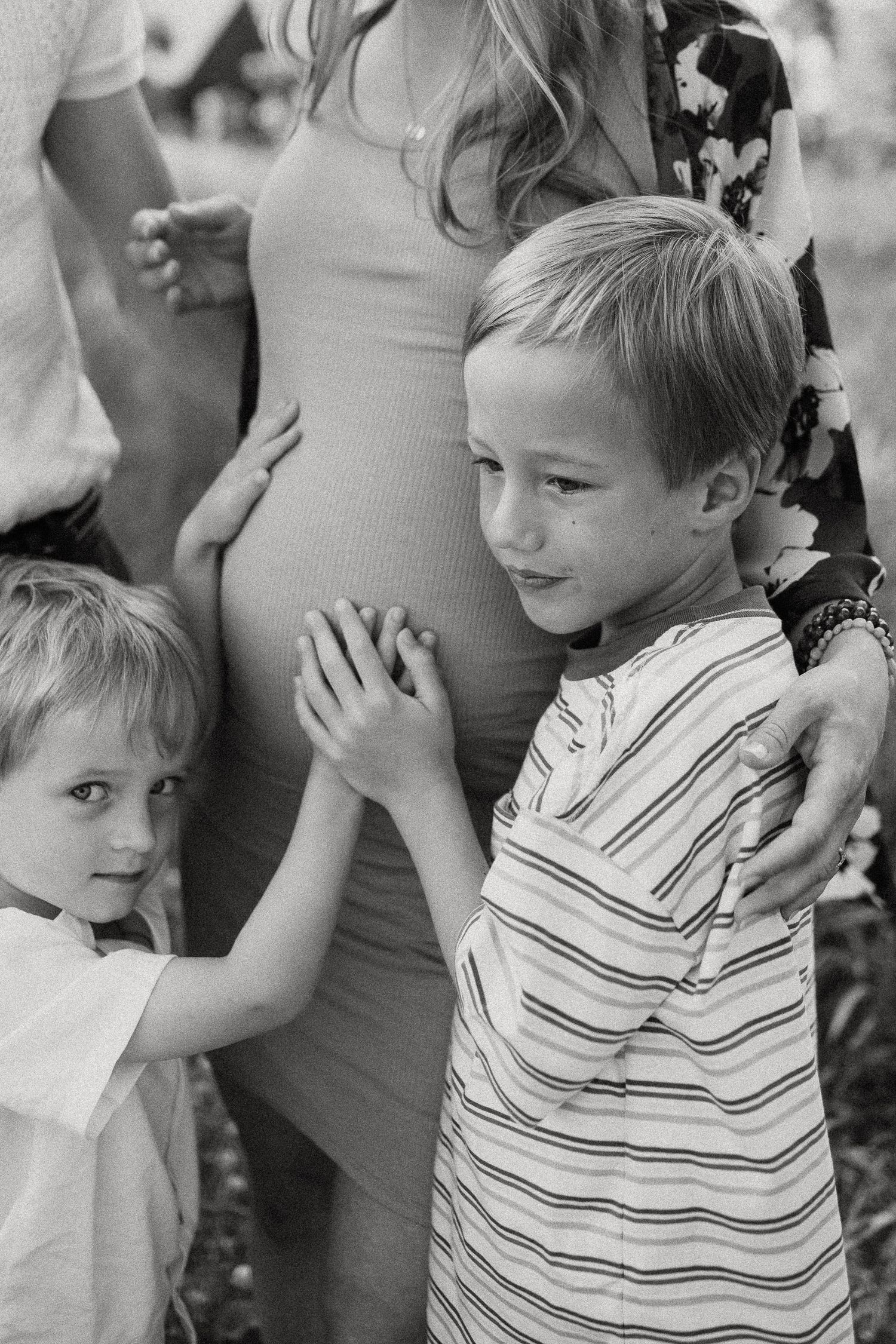 Two young boys tenderly touch their pregnant mothers belly in a black and white outdoor portrait.