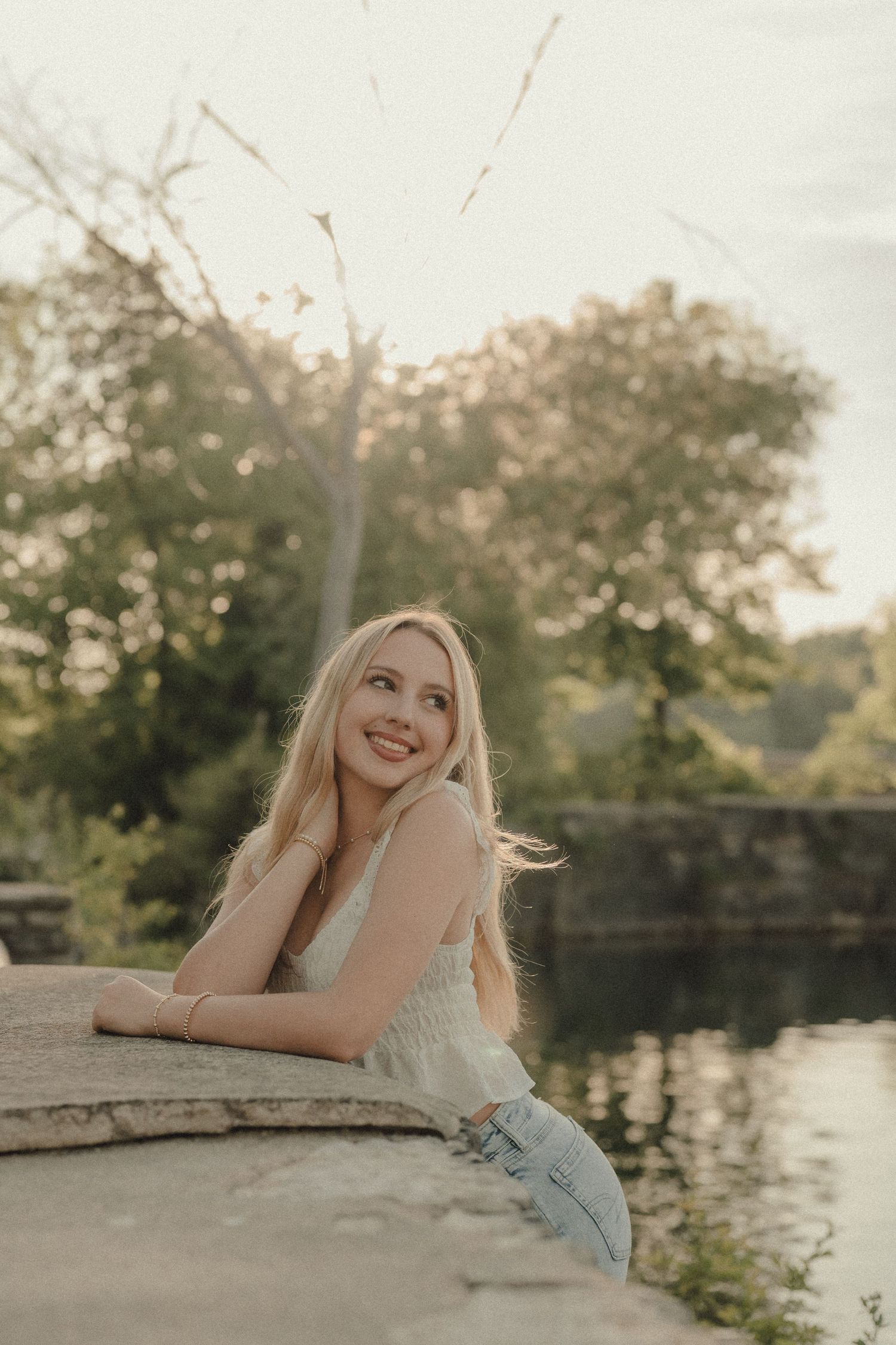 Smiling blonde woman leaning on stone wall by a serene river, surrounded by lush green trees in warm golden light.