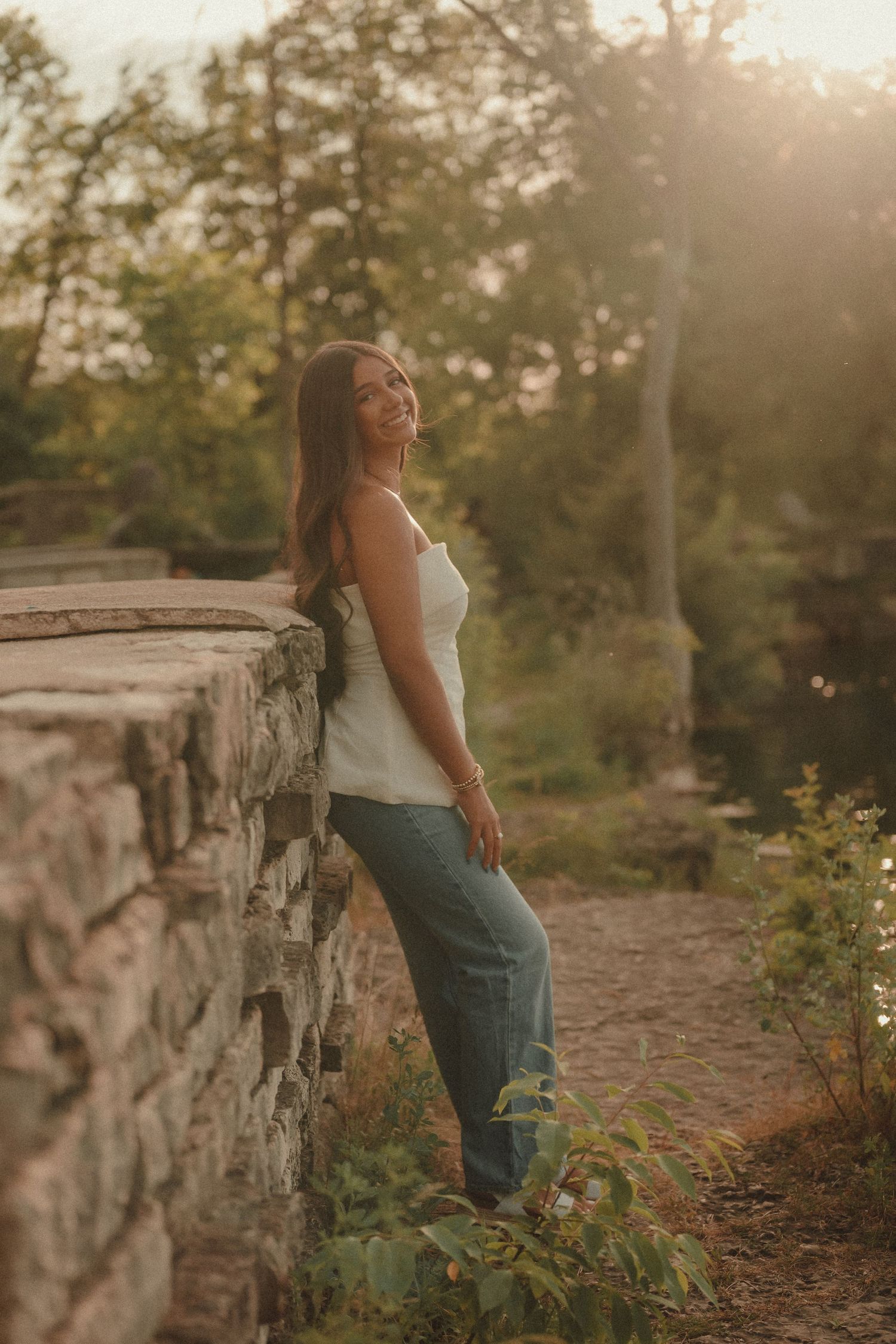Young woman in white tank top and jeans leaning against stone wall in golden hour sunlight outdoors.