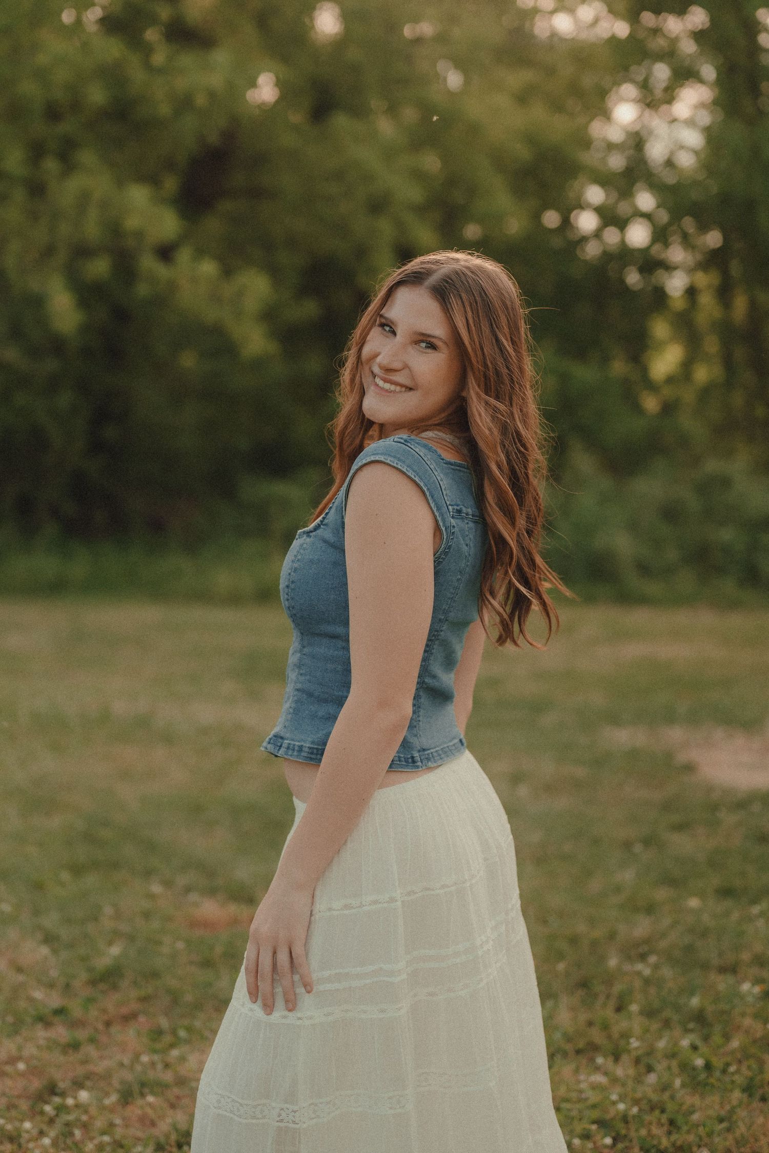 Young woman in denim vest and white tulle skirt smiling over shoulder outdoors.