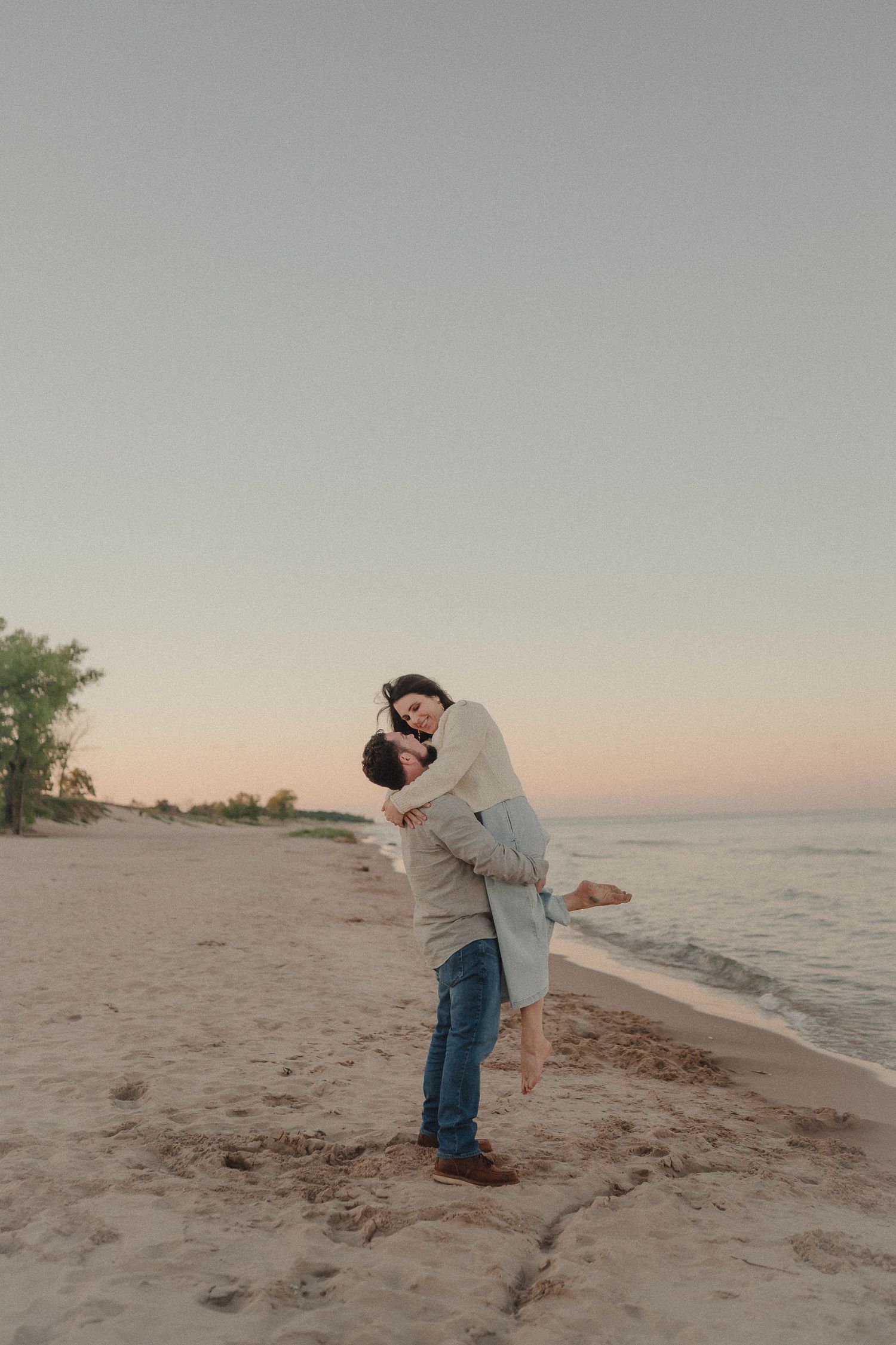 Man lifts woman joyfully on sandy beach during romantic golden hour sunset session.