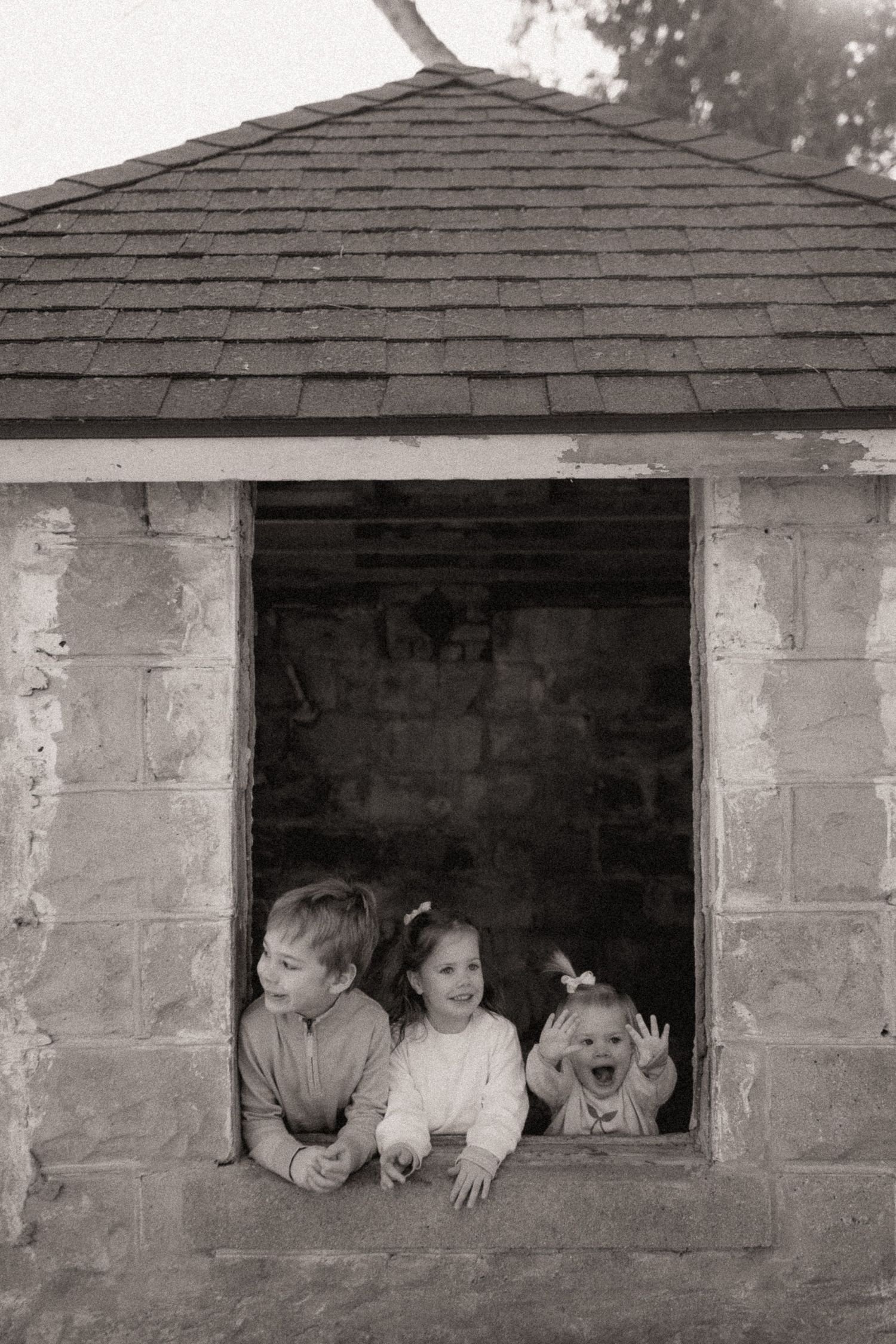 Children peek and play through a window opening in a concrete block wall in black and white photos.