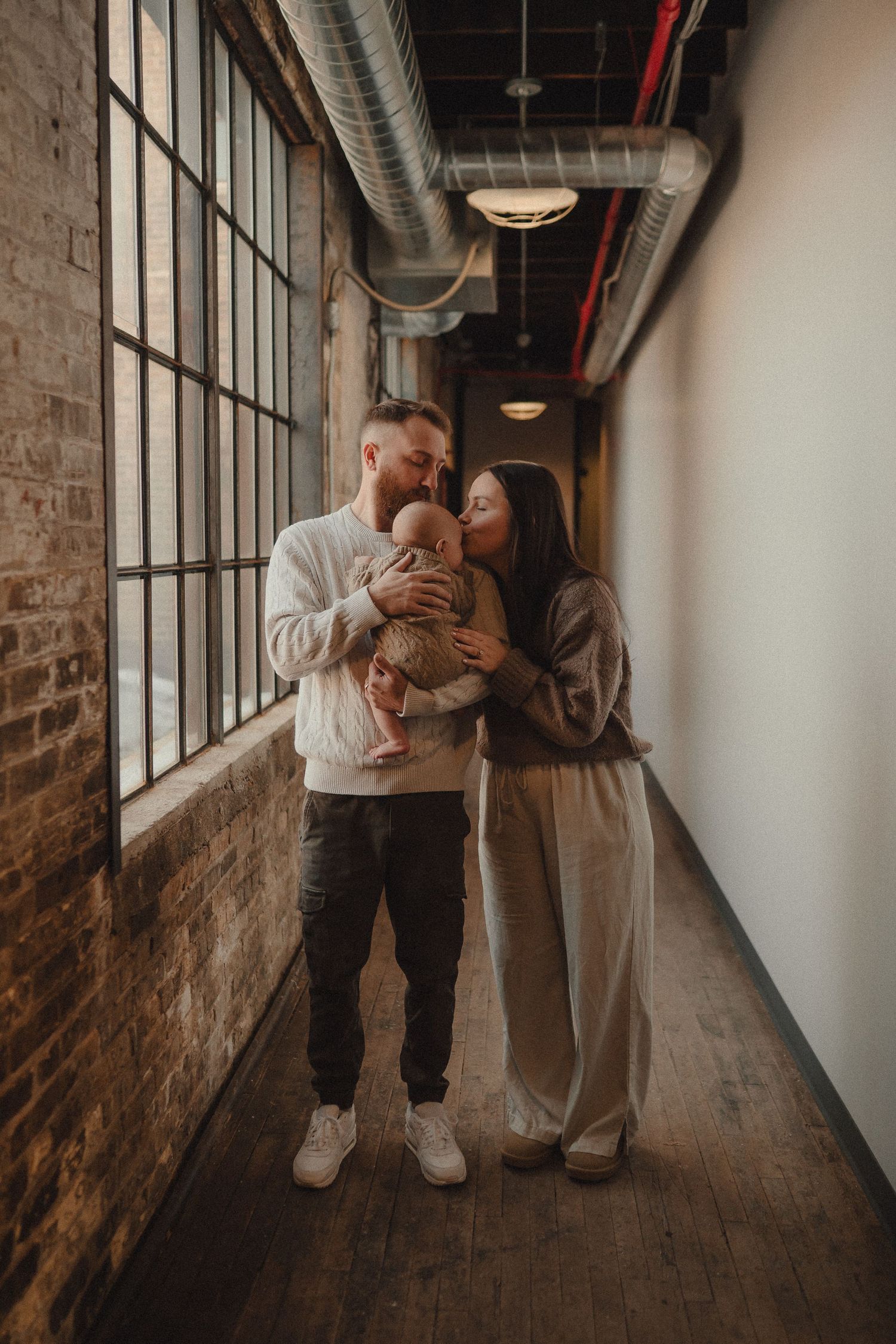 Tender family moments in an industrial hallway with large windows and exposed brick walls.