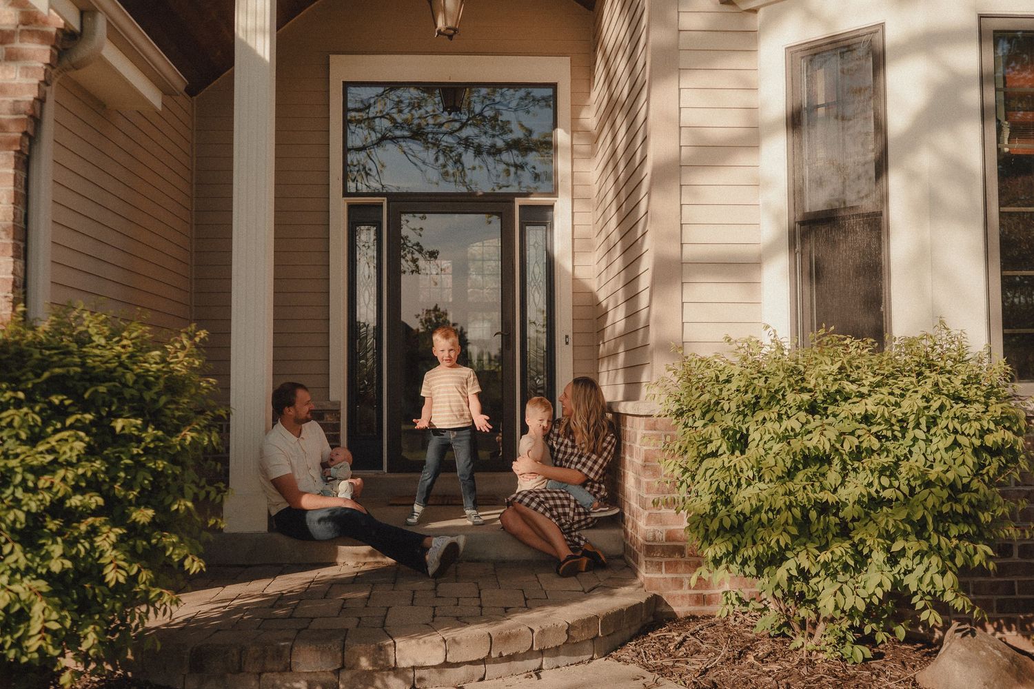 Family of four relaxing on front porch steps of a suburban home surrounded by lush greenery in warm sunlight.