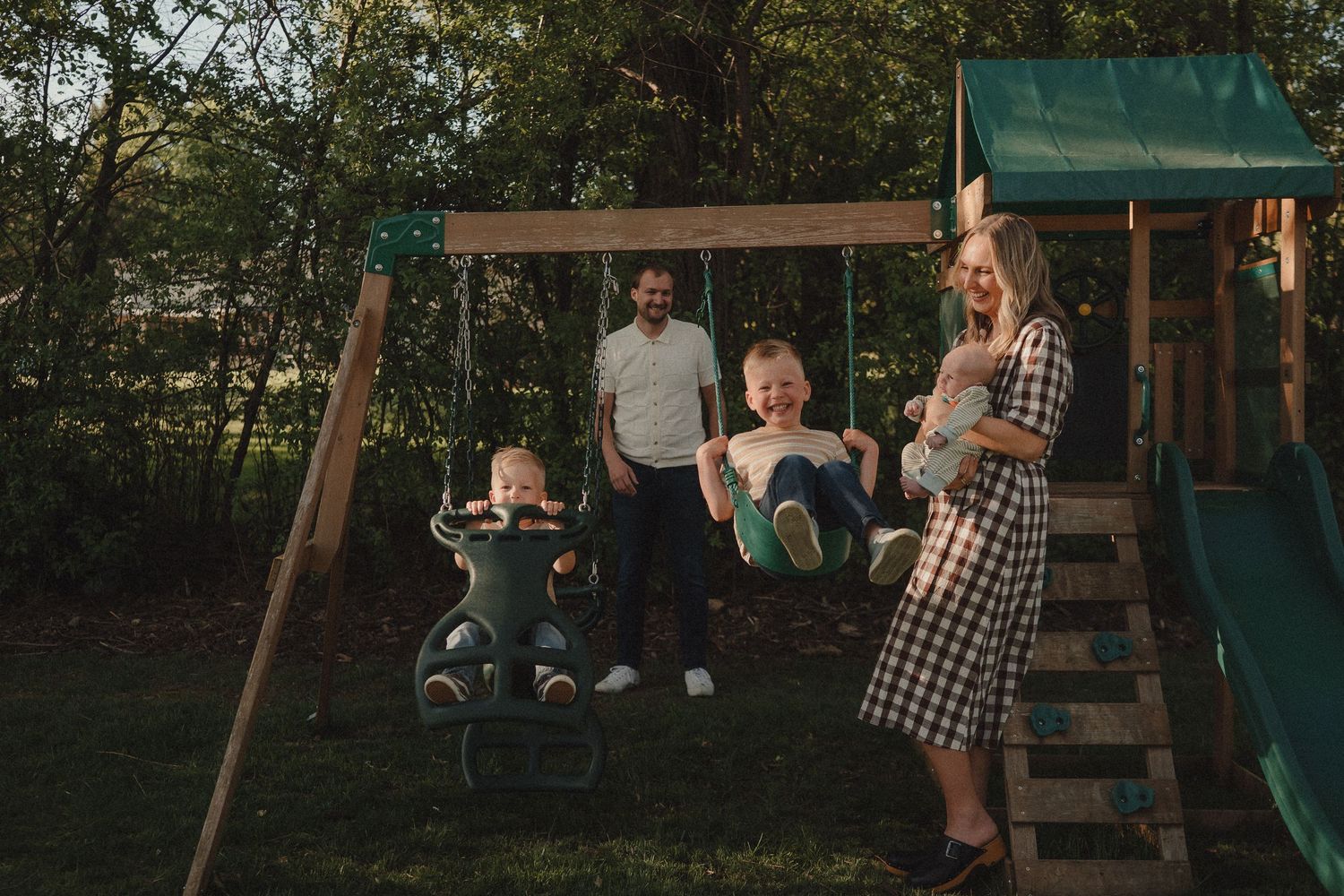 Family enjoying a backyard playset with children on swings while parents watch and smile on a sunny day.