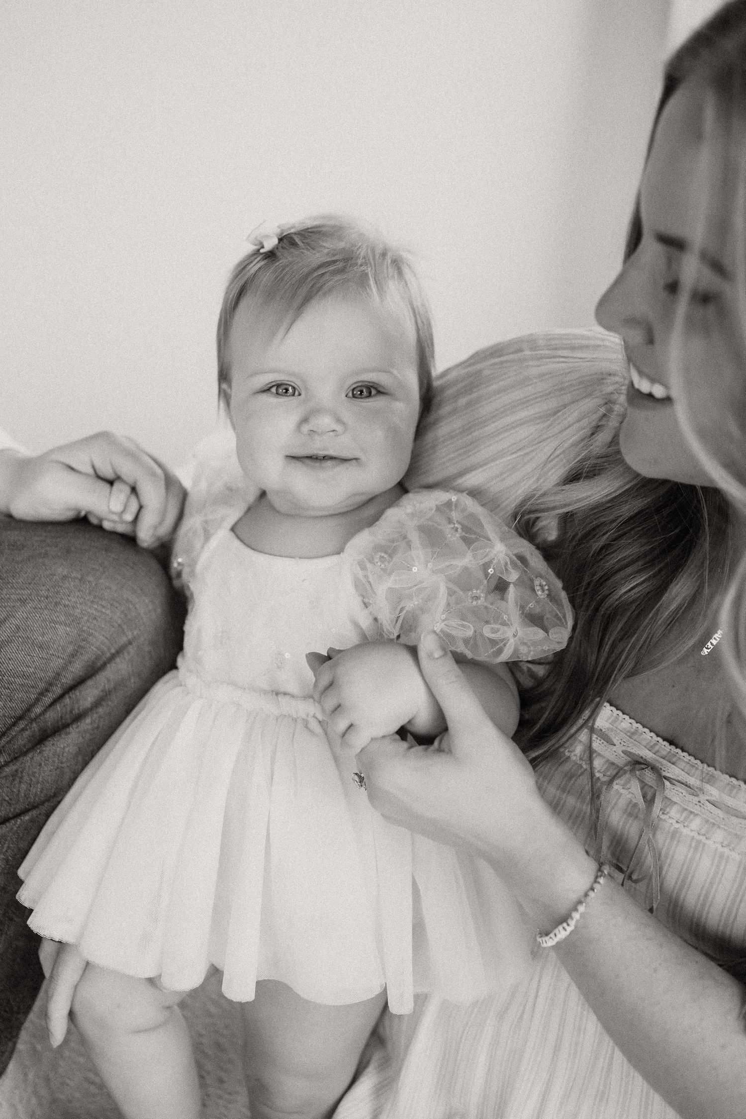 Black and white portrait of a smiling baby girl in a white tulle dress held by her parents.
