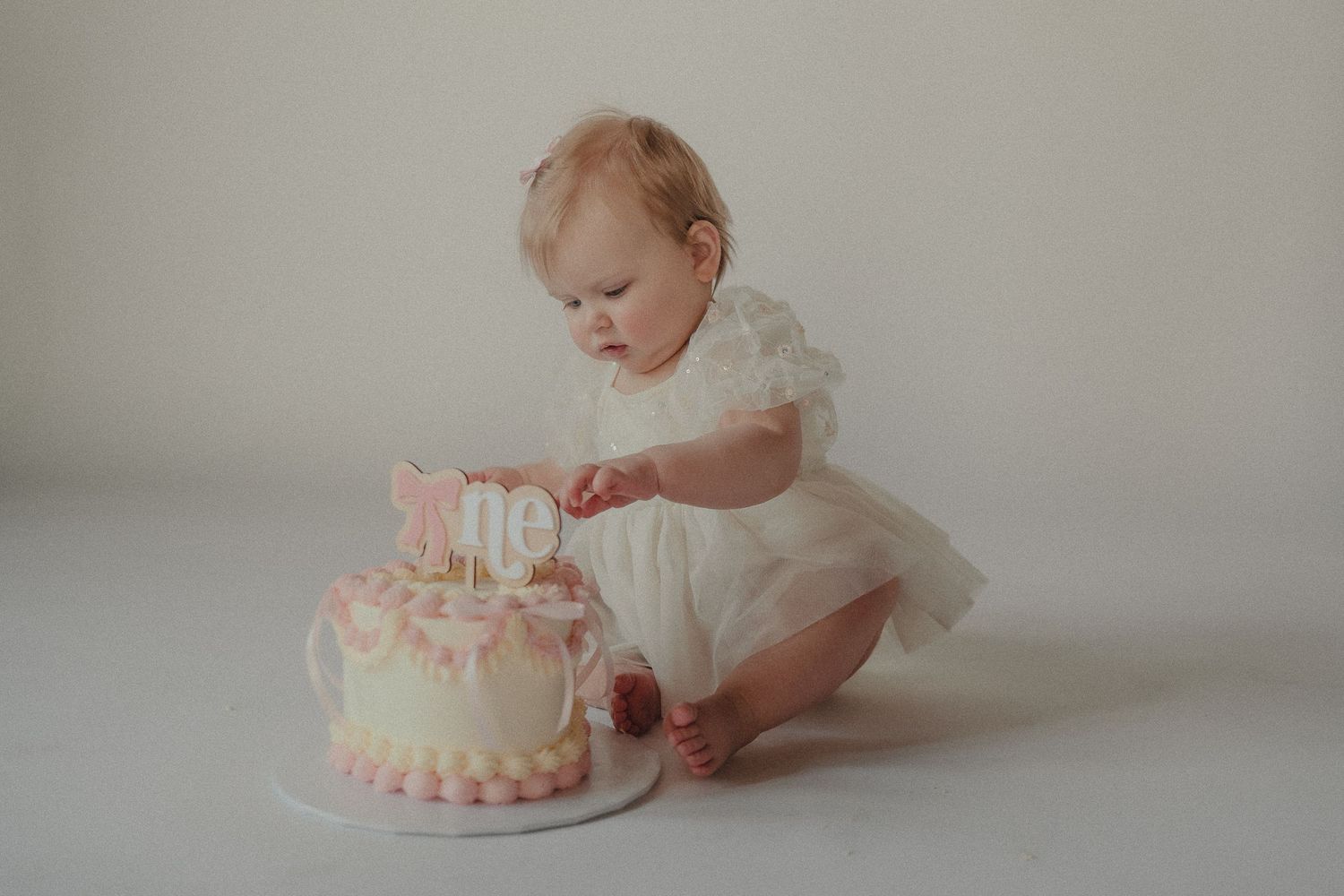 An adorable baby girl in a white dress reaches toward her pink and white first birthday smash cake.