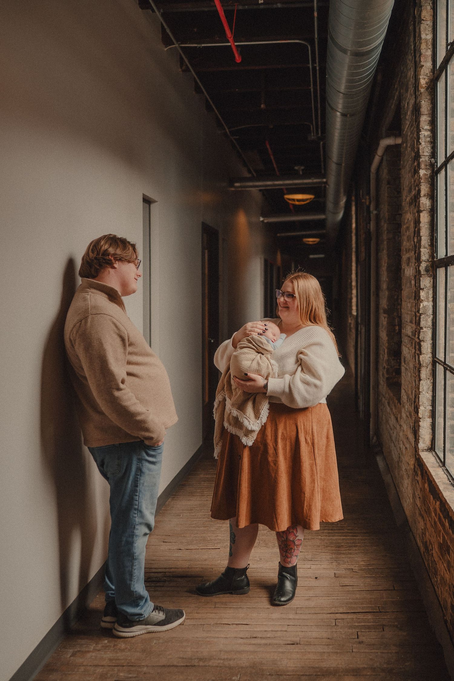 Couple in cozy sweaters stand in industrial hallway as woman cradles newborn baby wrapped in white blanket.