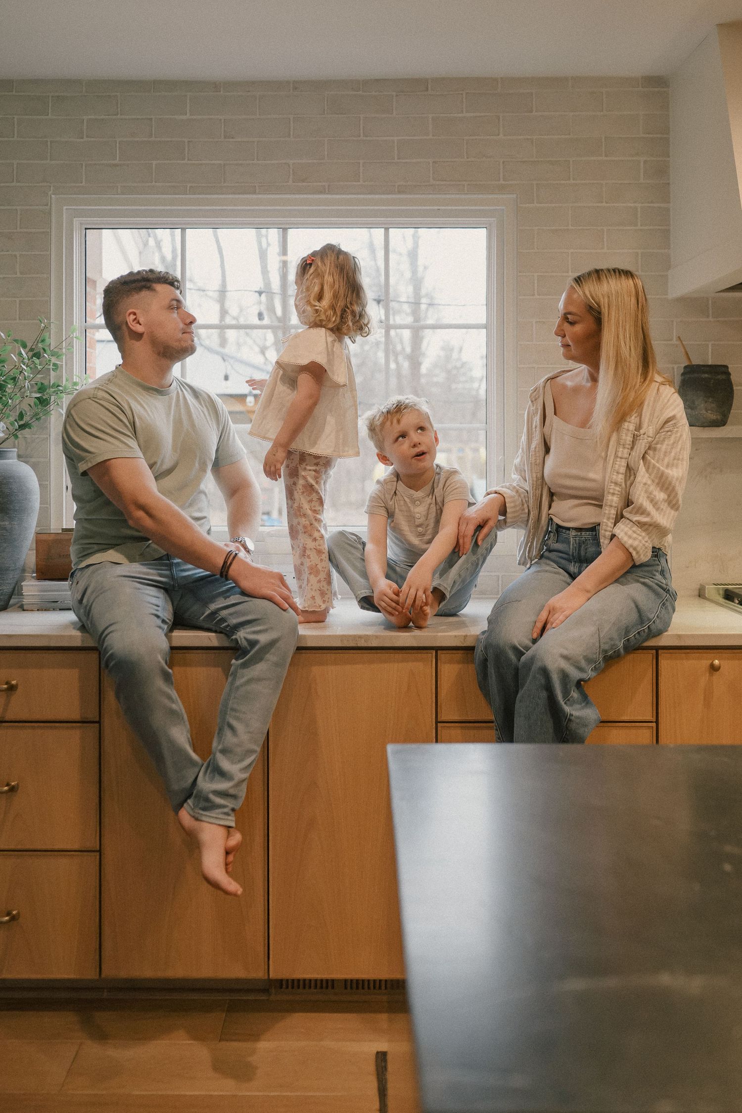 Family of four sitting on a kitchen counter by a large window in a warm modern kitchen.