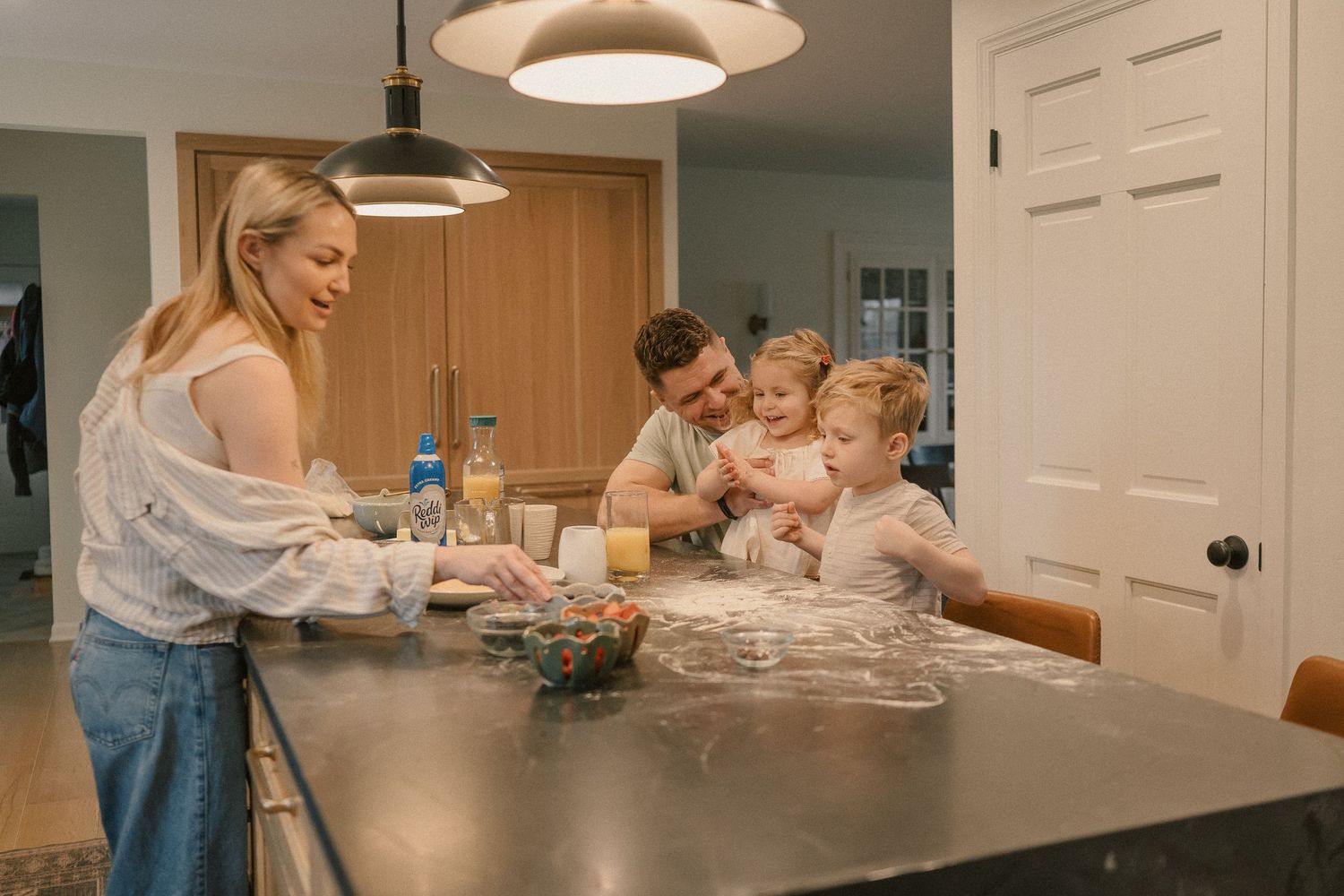 Family baking together at kitchen island, parents and two young children playing with flour dough at home.