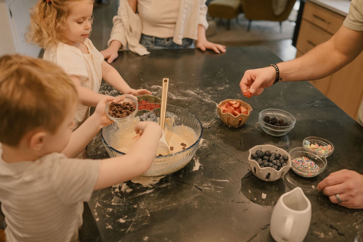 Family adding fresh fruits and chocolate chips to a mixing bowl while baking together in a modern kitchen.