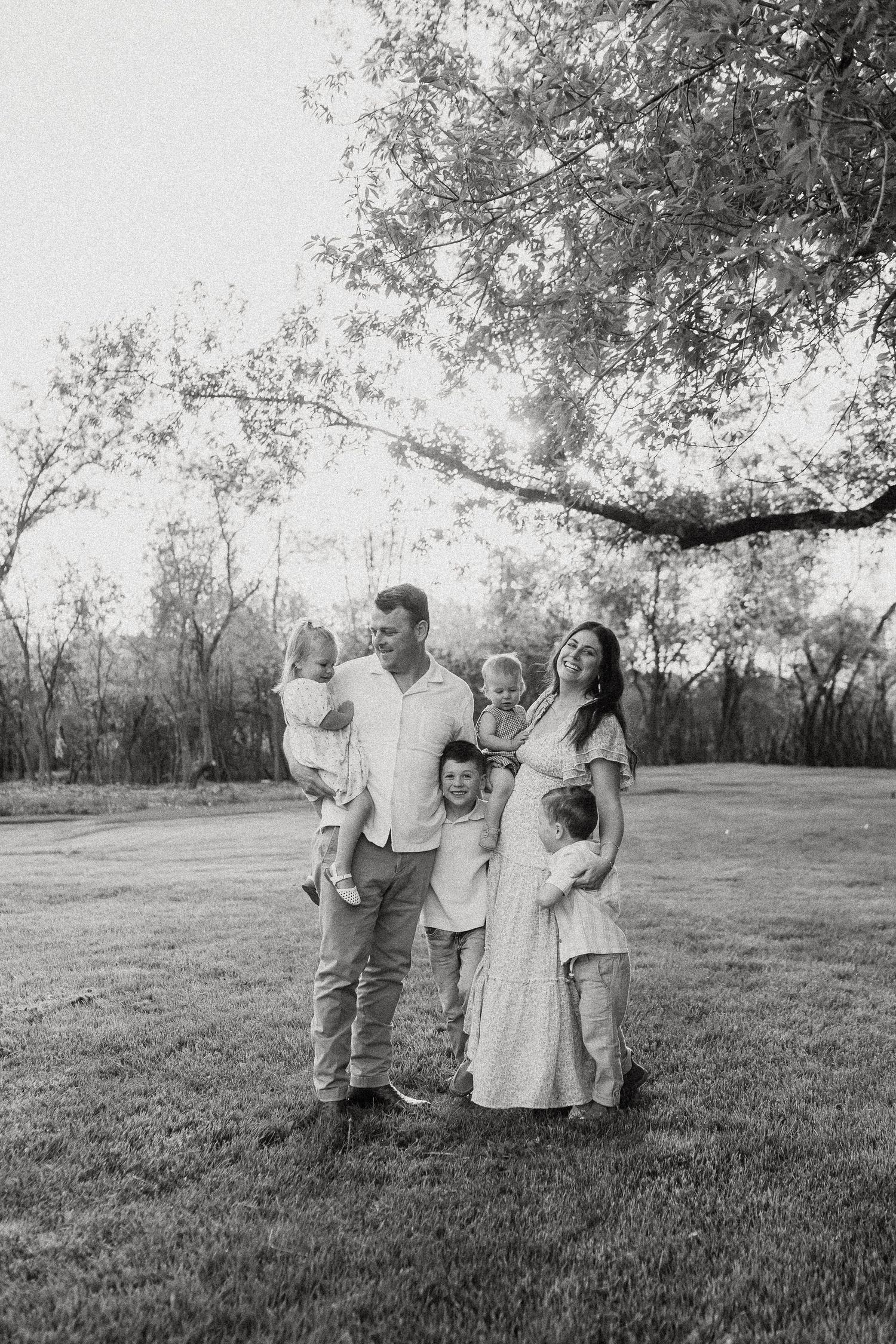 Black and white family portrait of parents with four young children standing under trees in park.