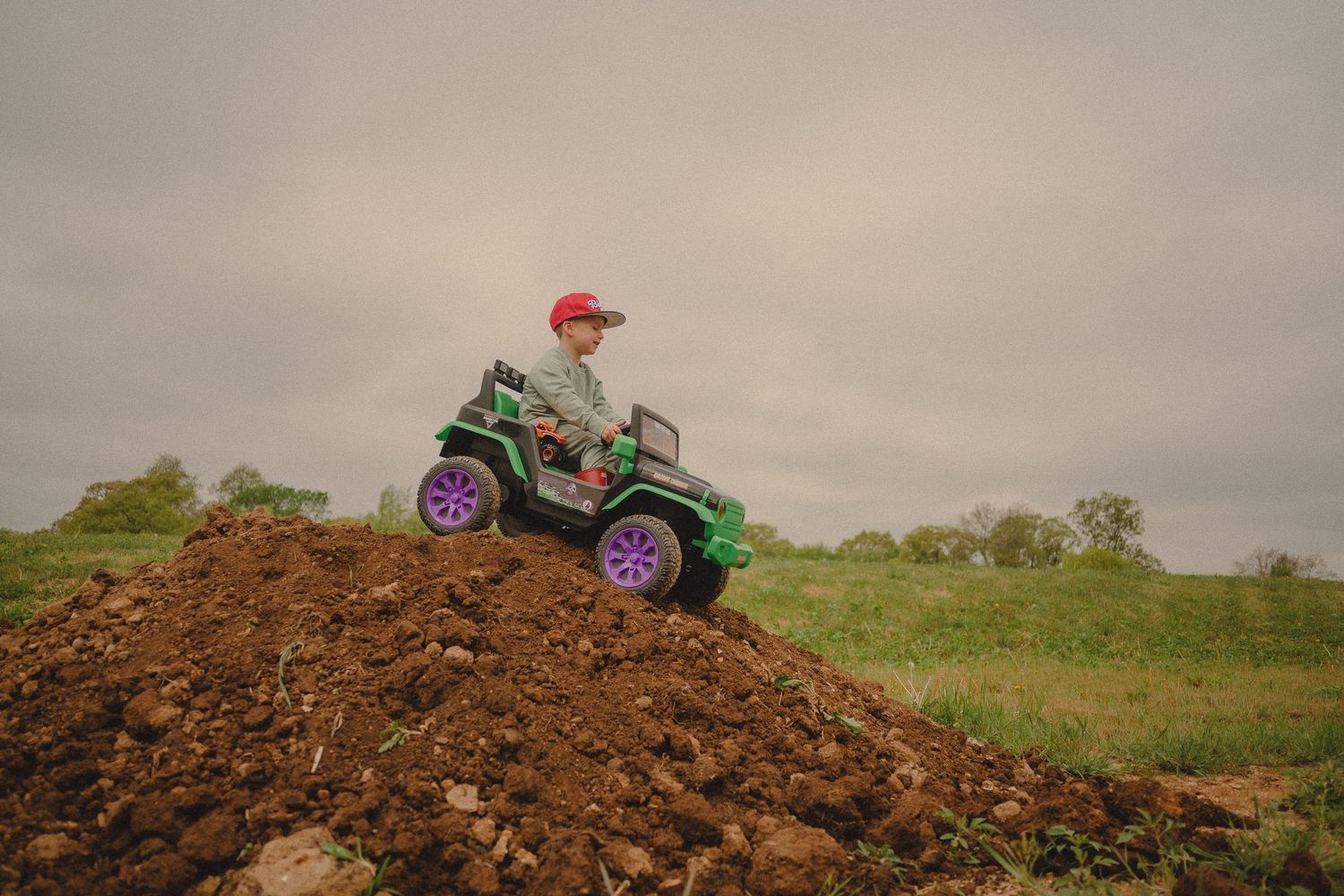 Young boy in red cap rides green toy ATV on dirt mound in overcast outdoor setting.