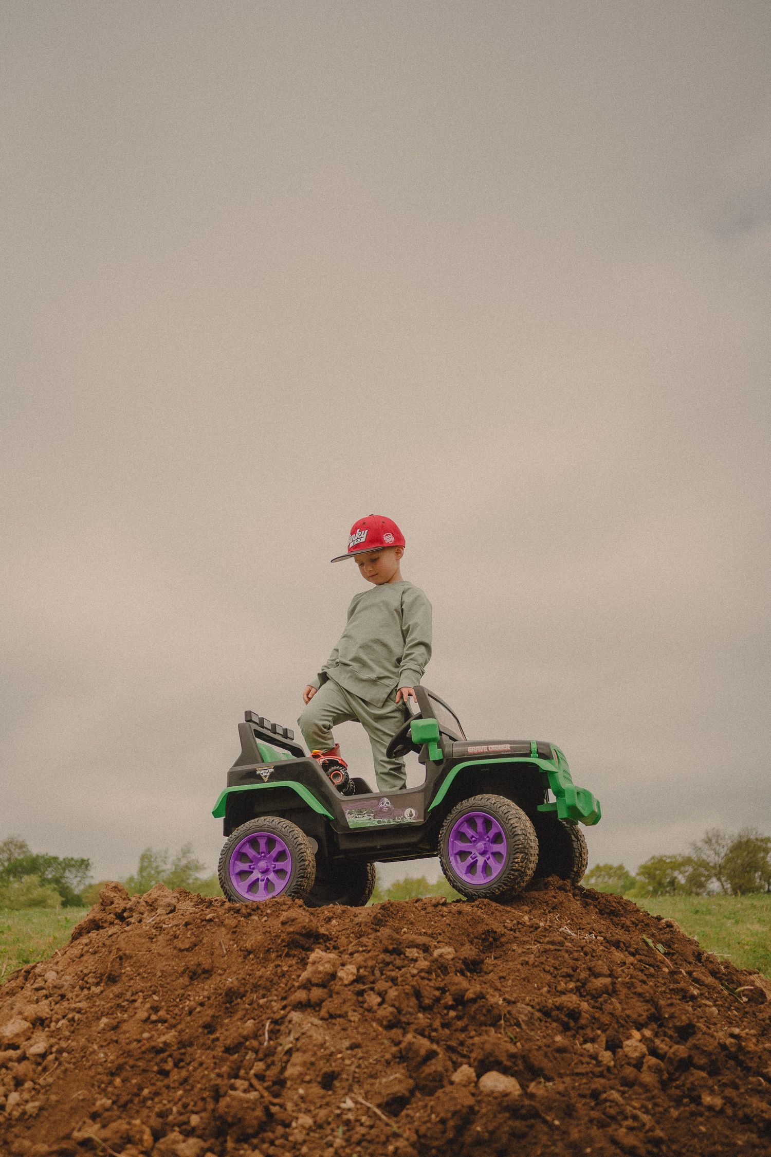 Young boy in red cap rides a green electric toy jeep atop a dirt hill outdoors.