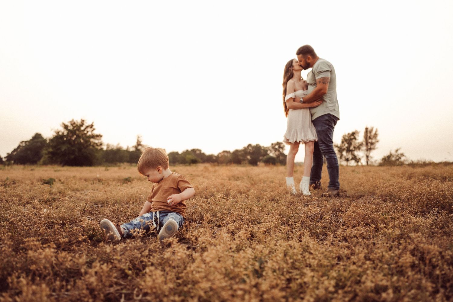 A romantic sunset moment in a golden field as a couple embraces while a toddler plays nearby.