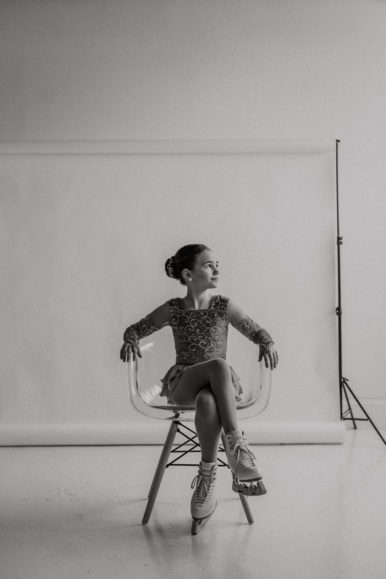 Figure skater in sparkly costume poses on clear chair in dramatic black and white studio portrait.